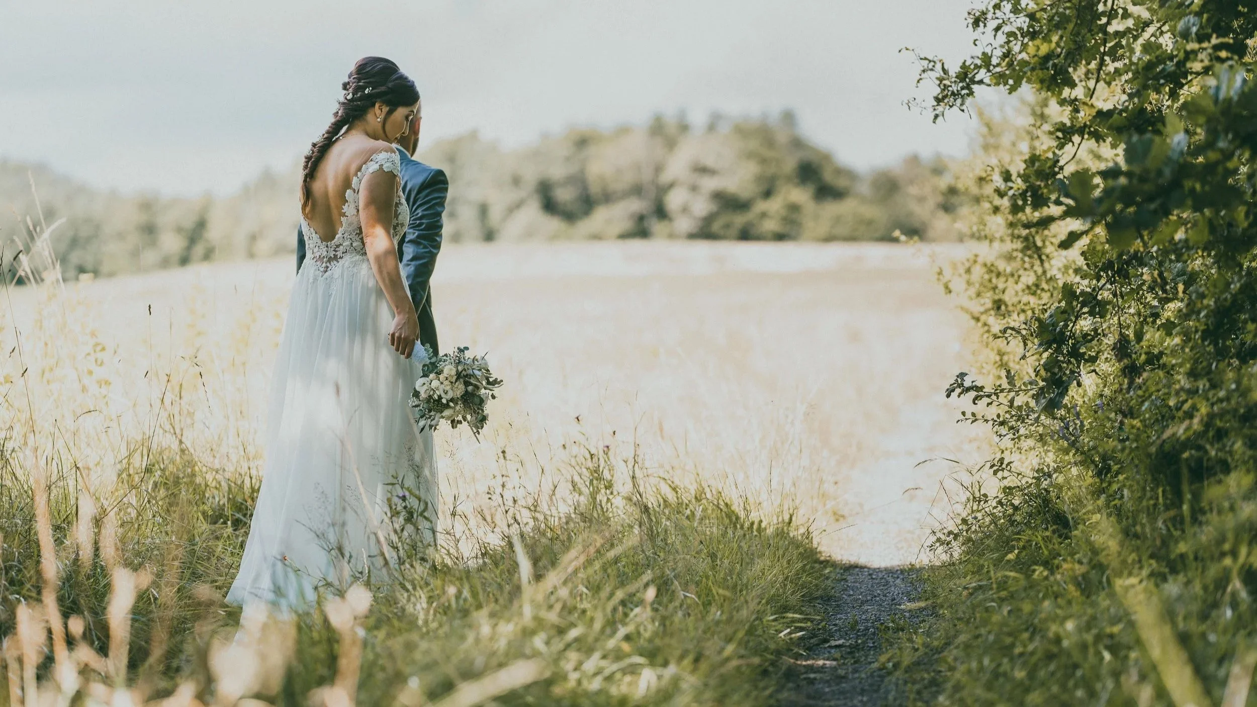 Ein Brautpaar steht im Grünen, das Brautkleid ist weiß mit Spitze, die Szene ist im Freien.
