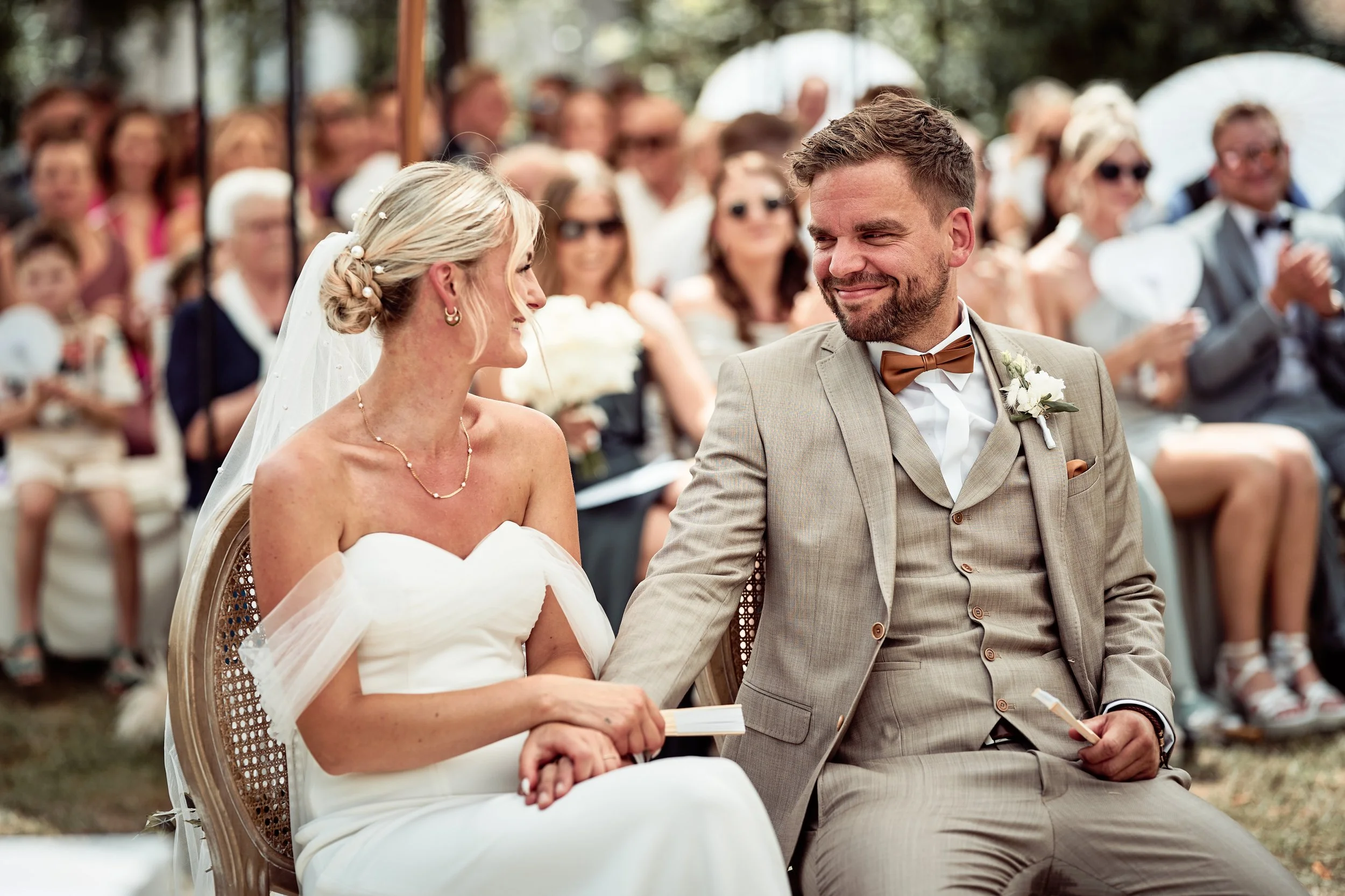 Braut und Bräutigam bei ihrer Hochzeit, Sitzend und Händchen haltend, lächelnd, im Hintergrund Hochzeitsgäste, draußen, sonniges Wetter.