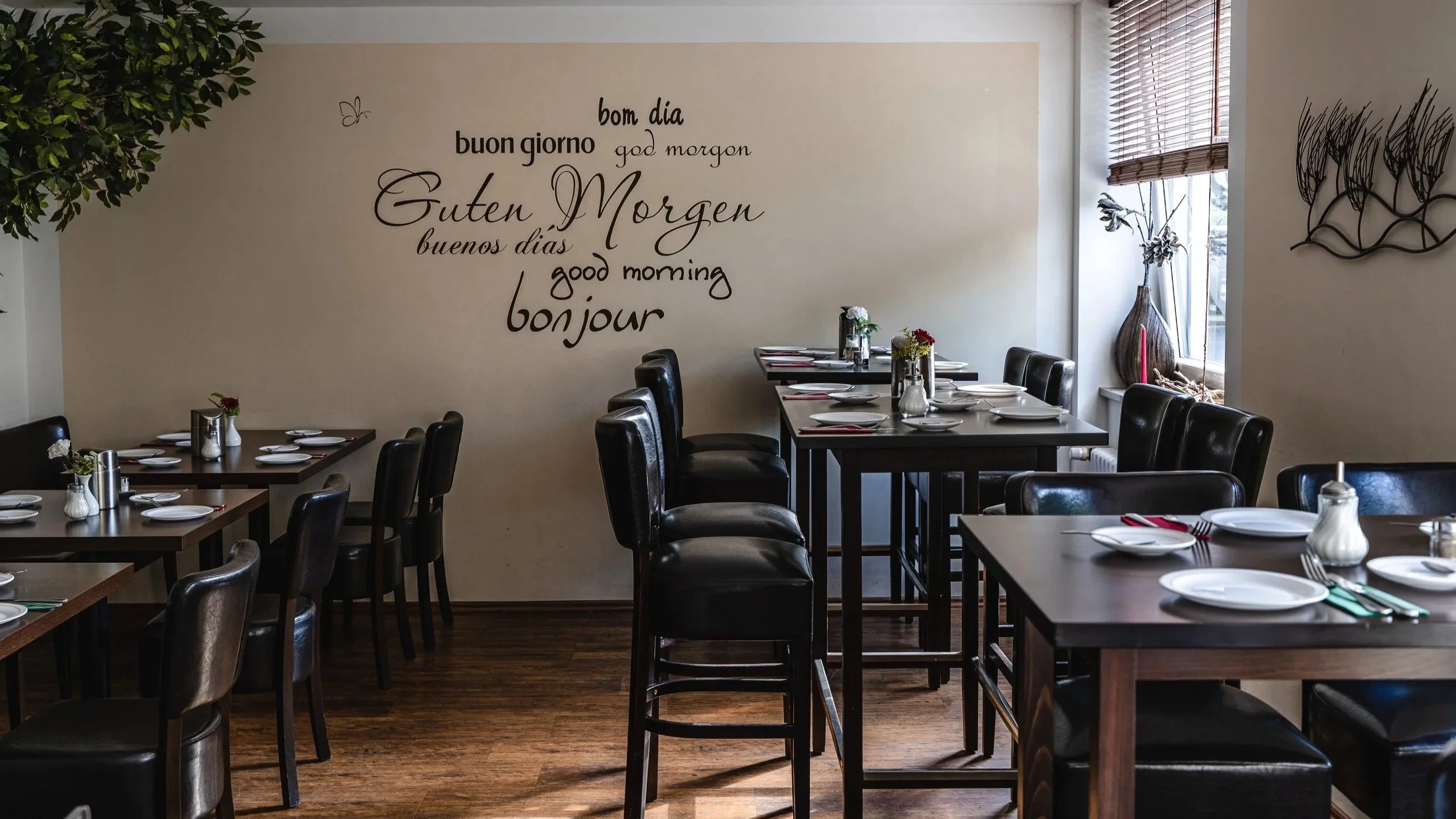 Ein leerer Speiseraum in einem Restaurant mit dunkelbraunen Tischen und schwarzen Stühlen, Dekorationen auf den Tischen, und einer weißen Wand mit mehrsprachigen Begrüßungswörtern für Guten Morgen. Es sind Fenster mit Rollläden, und Wanddekorationen sind sichtbar.