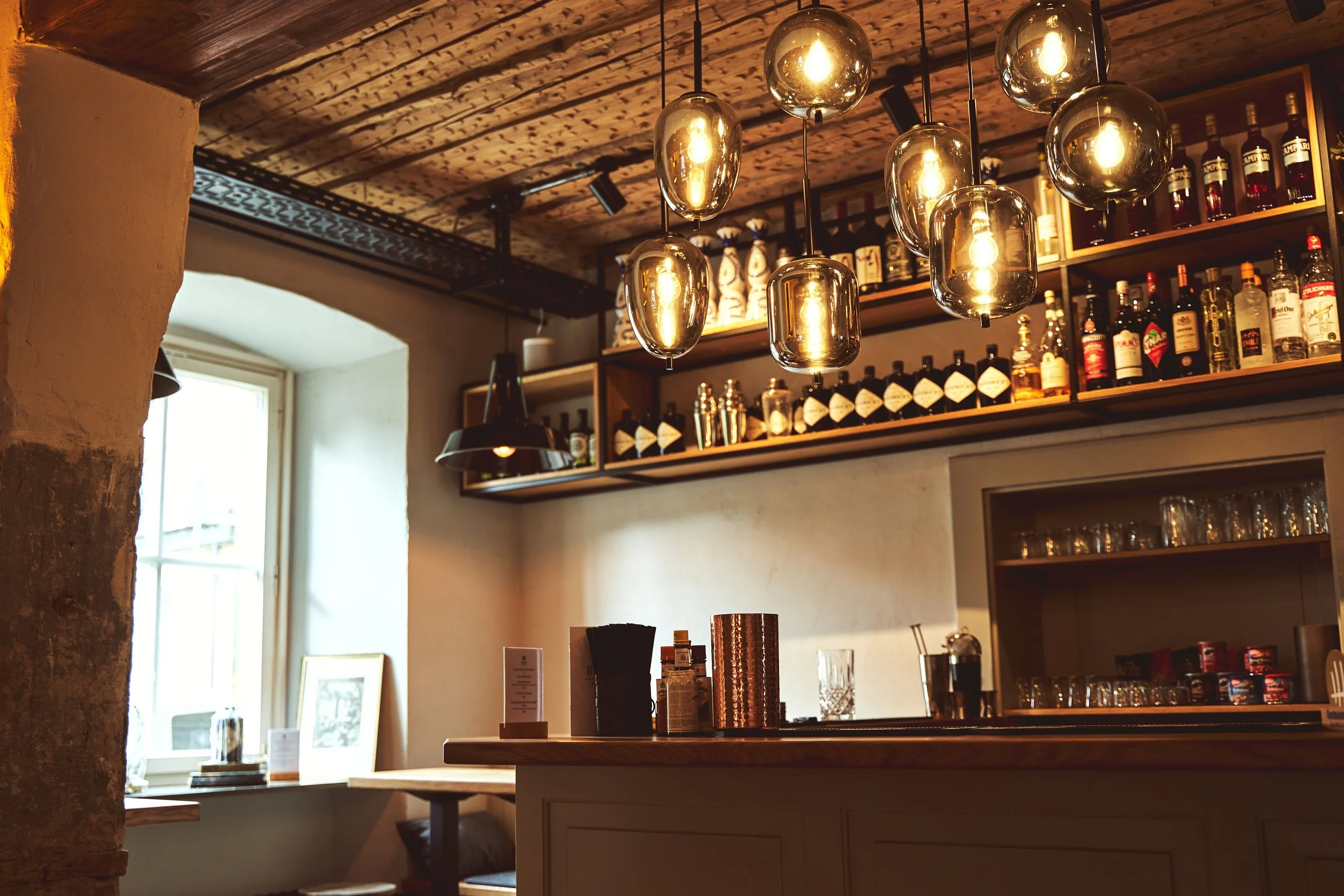 Innenansicht einer Bar mit hölzerner Decke, kelchförmigen Hängelampen und einer Regalkette mit Spirituosenflaschen