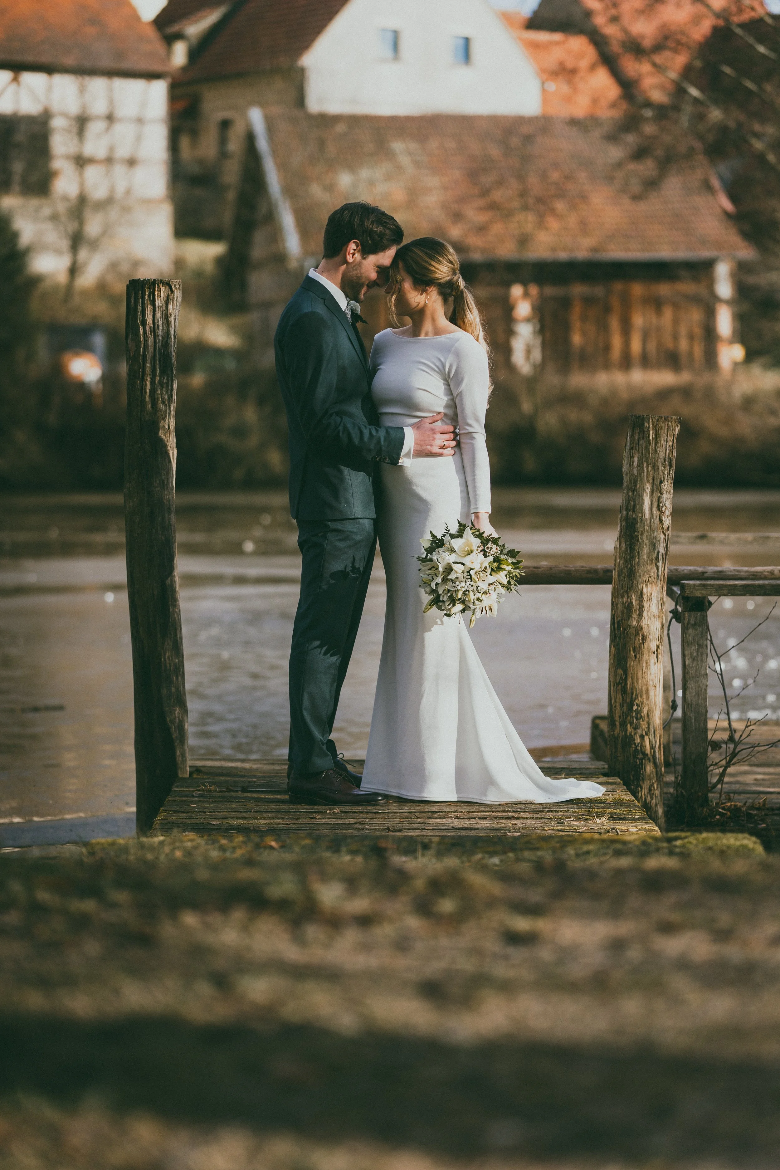 Ein Brautpaar steht auf einem Steg am Wasser, umgeben von alten Holzbalken, bei Sonnenuntergang, im Hintergrund alte Häuser in einem ländlichen Umfeld.