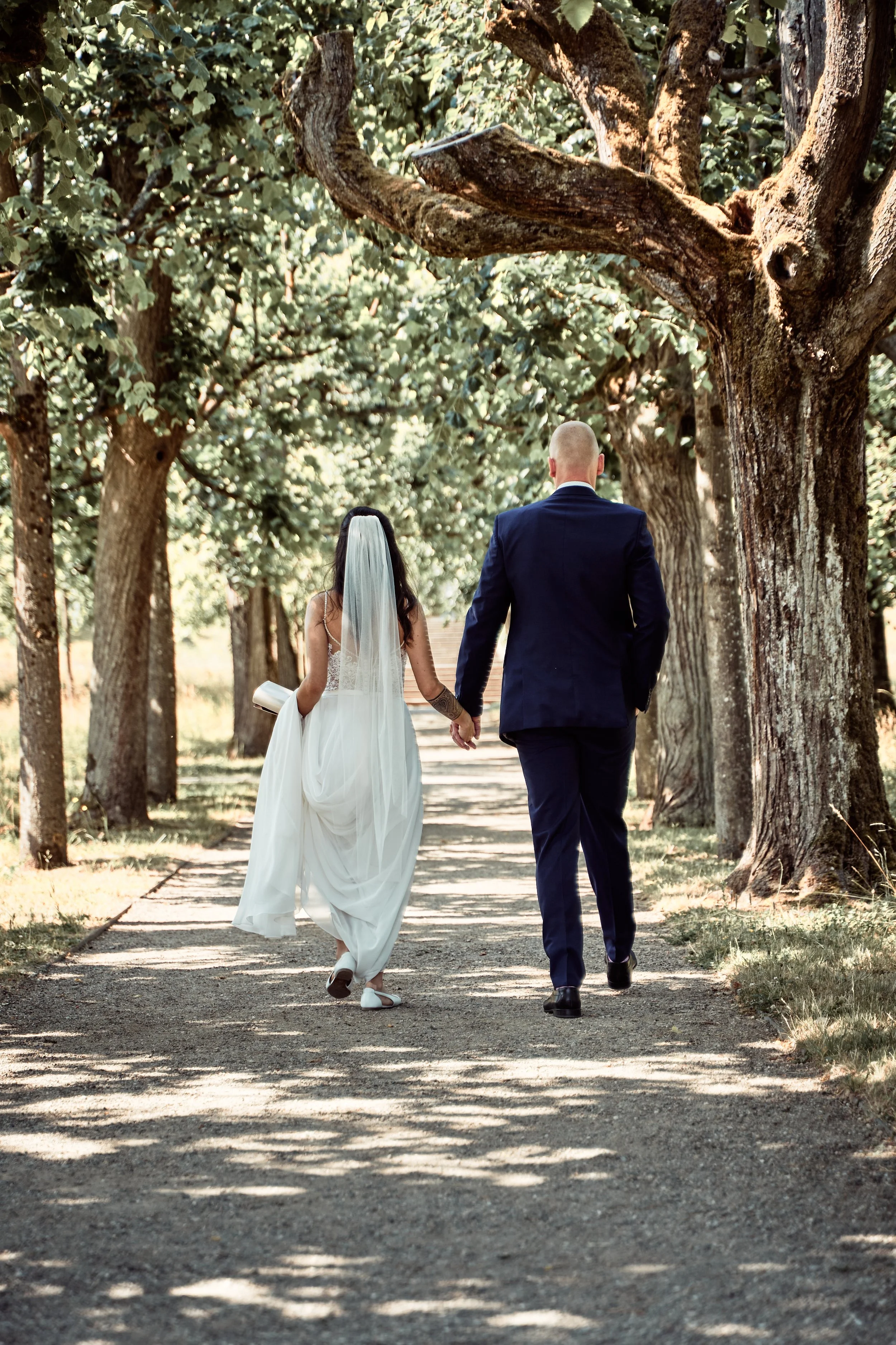 Ein Brautpaar läuft Hand in Hand auf einem Baumweg bei Sonnenlicht.