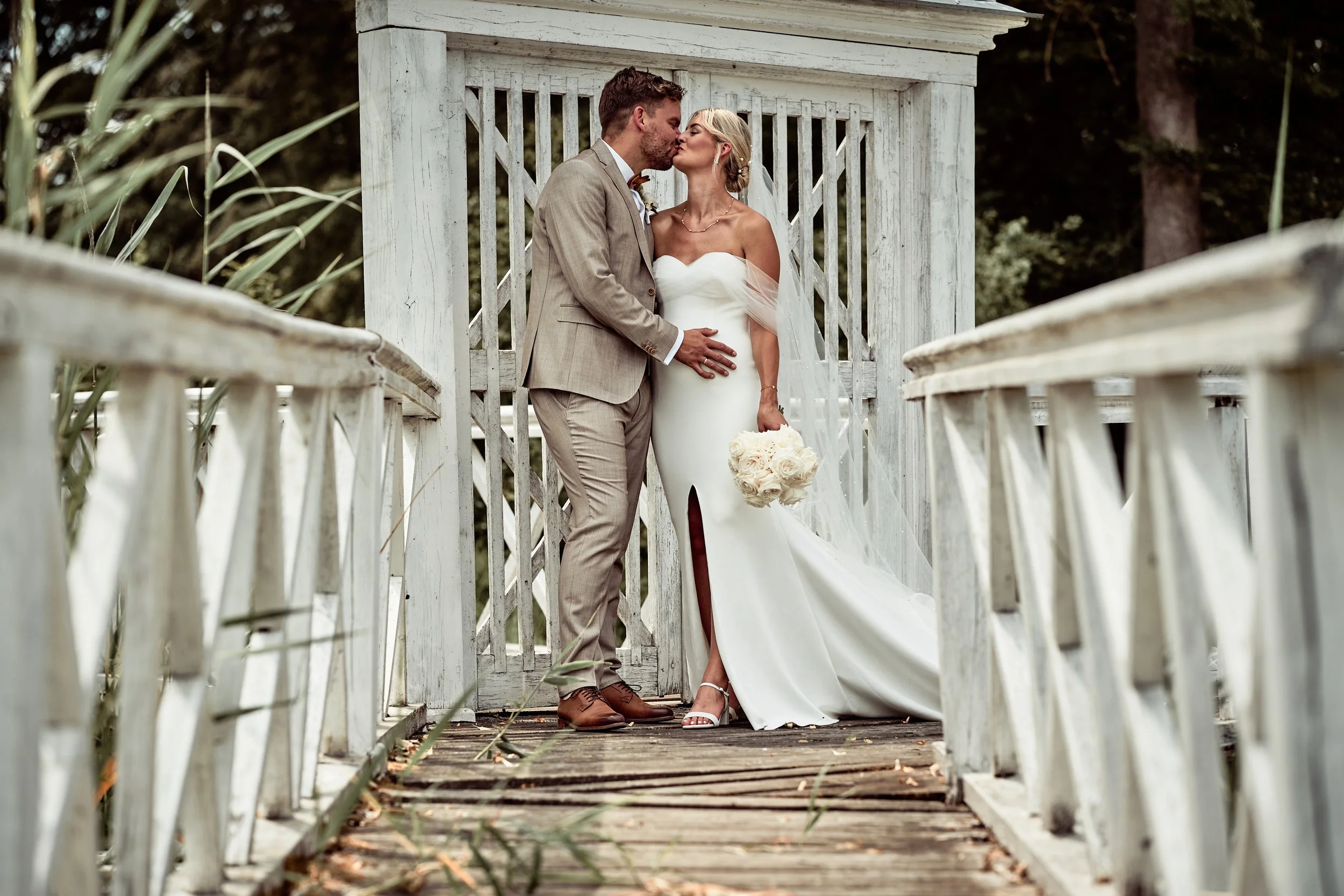 Ein frisch vermähltes Paar küsst sich auf einer kleinen Brücke im Freien, die Braut trägt ein weißes Hochzeitskleid und hält einen Blumenstrauß, der Bräutigam trägt einen beige Anzug.