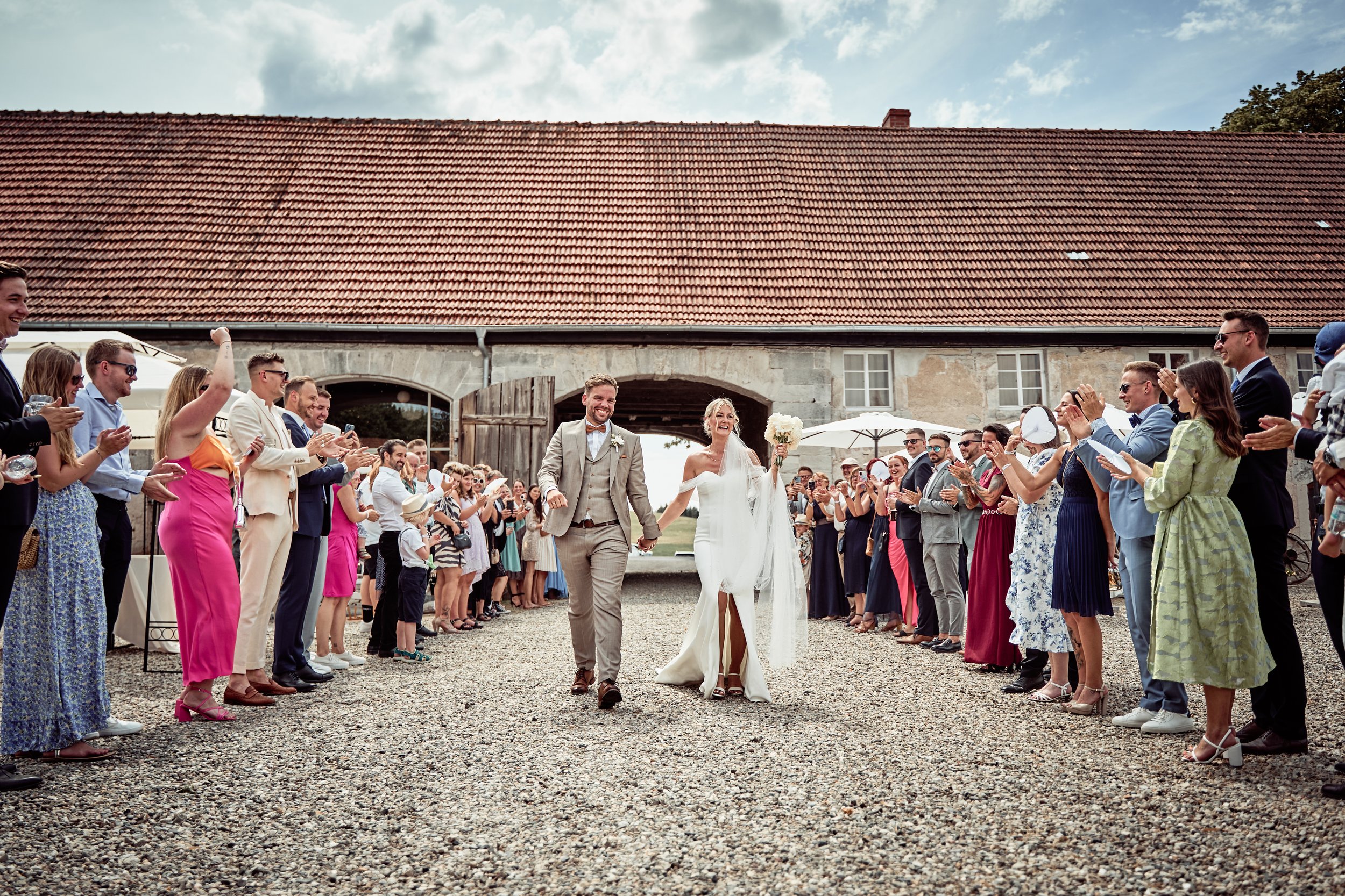 Ein Brautpaar geht Hand in Hand durch eine Menschenmenge bei einer Hochzeit im Freien, umgeben von Gästen, die applaudieren und feiern.