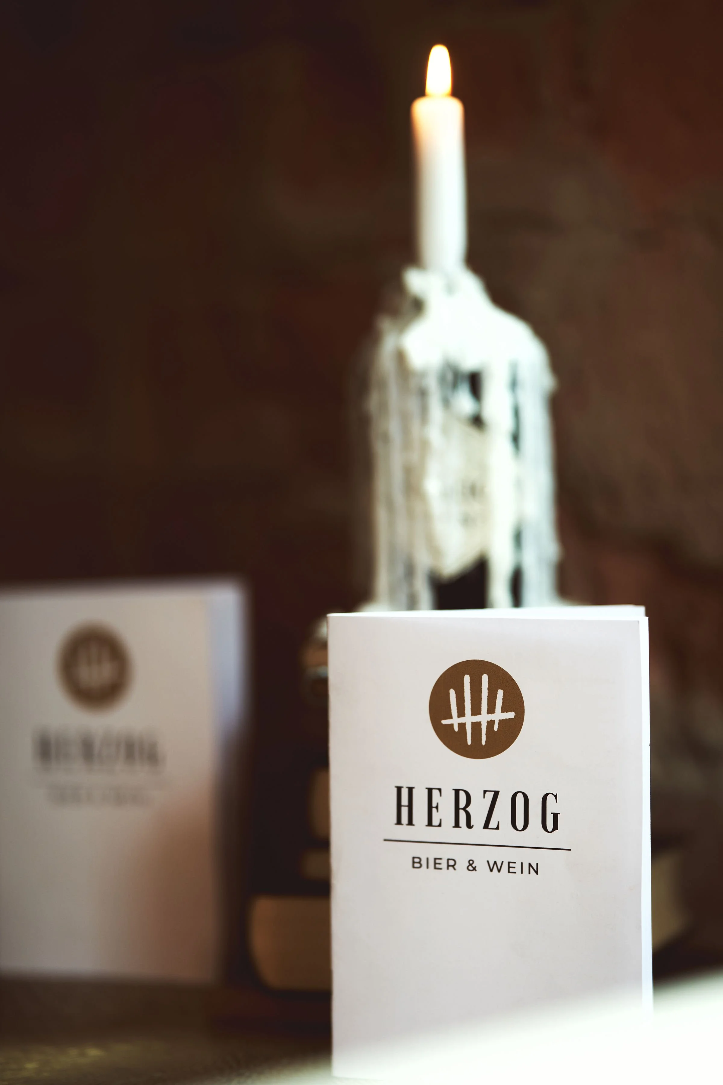 Eine weiße Speisekarte mit dem Schriftzug 'HERZOG BIER & WEIN' und einem Logo, im Hintergrund eine brennende Kerze in einer dekorativen Halterung.