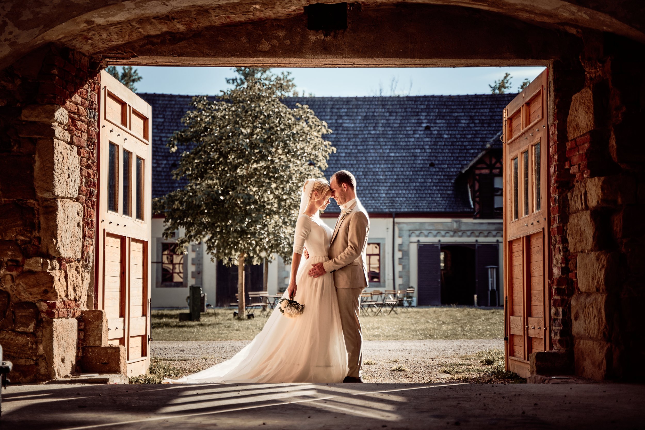 Ein Hochzeitsfoto zeigt ein Brautpaar, das sich an einem Tor umarmt, im Hintergrund ein Baum, alte Gebäude und eine klarer Himmel.