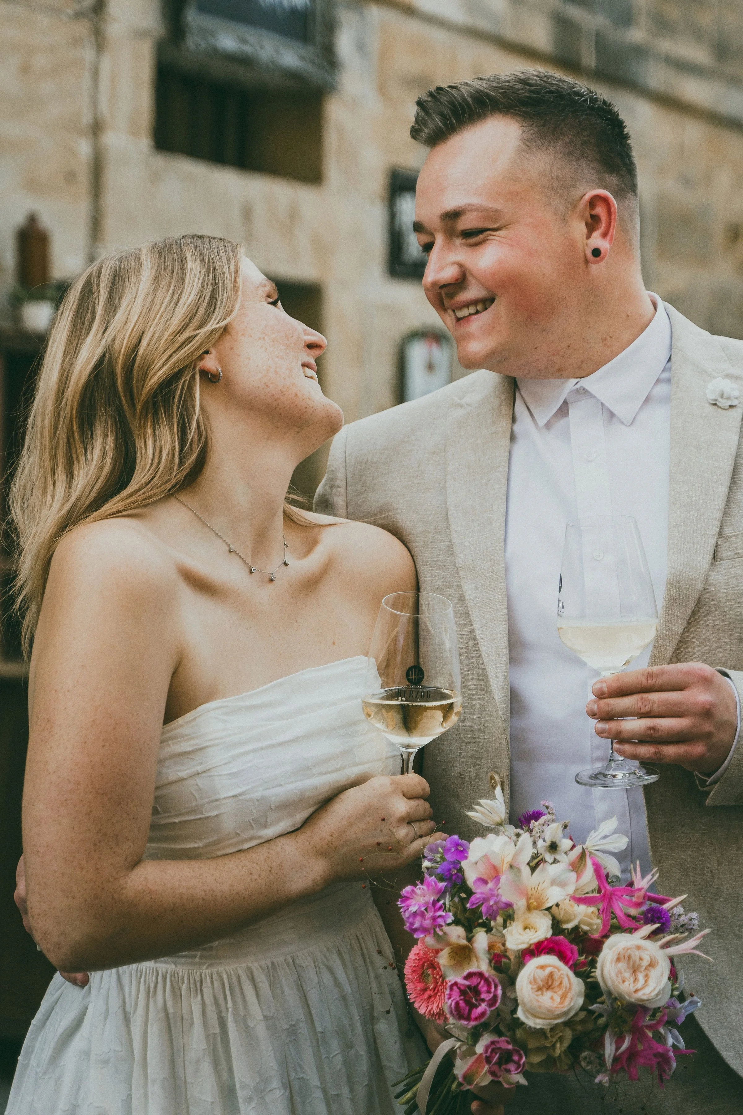 Ein glückliches Brautpaar bei einer Feier, beide halten Weingläser und lächeln sich an, die Braut hält einen Blumenstrauß.