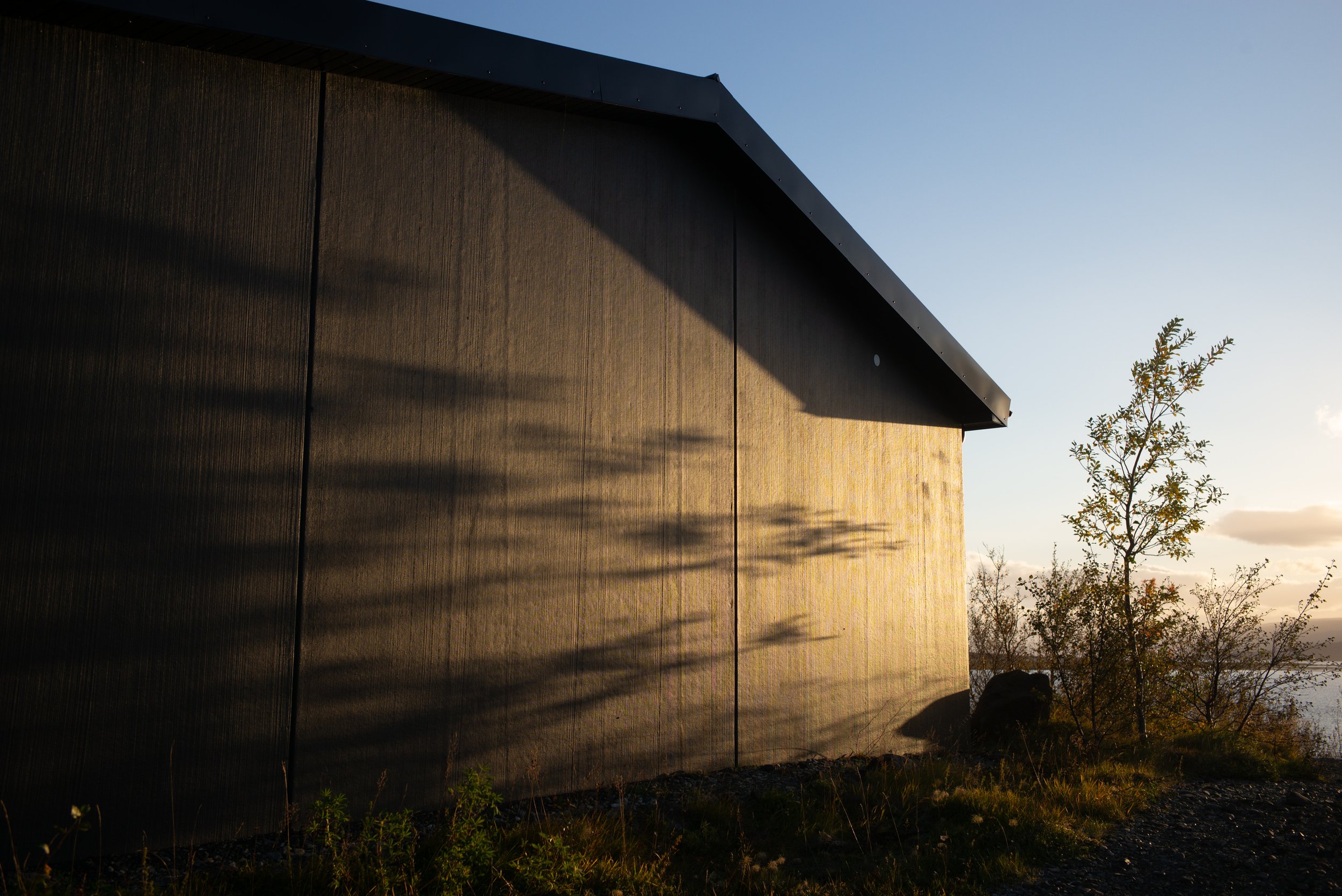 Ein dunkles Holzhaus bei Sonnenuntergang, das einen Schatten an die Wand projiziert, neben einer kleinen Baum auf einer Grasthe rippe.