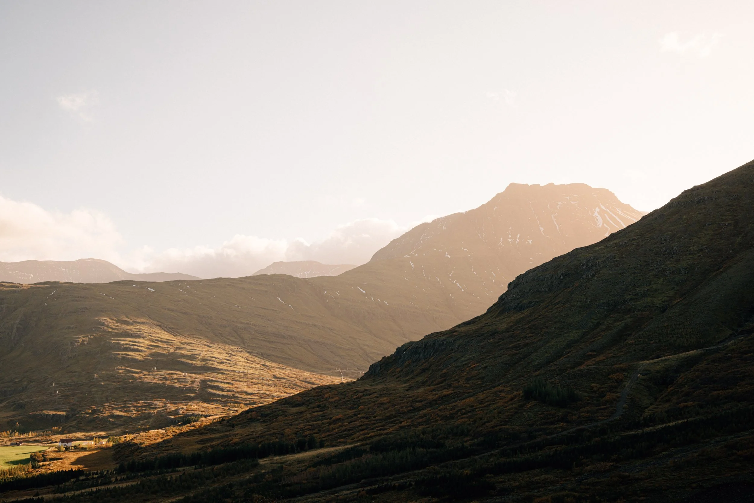 Berglandschaft mit sonnenbeschienenen Hügeln und einem hohen Gipfel im Hintergrund, unter einem Himmel mit wenigen Wolken.
