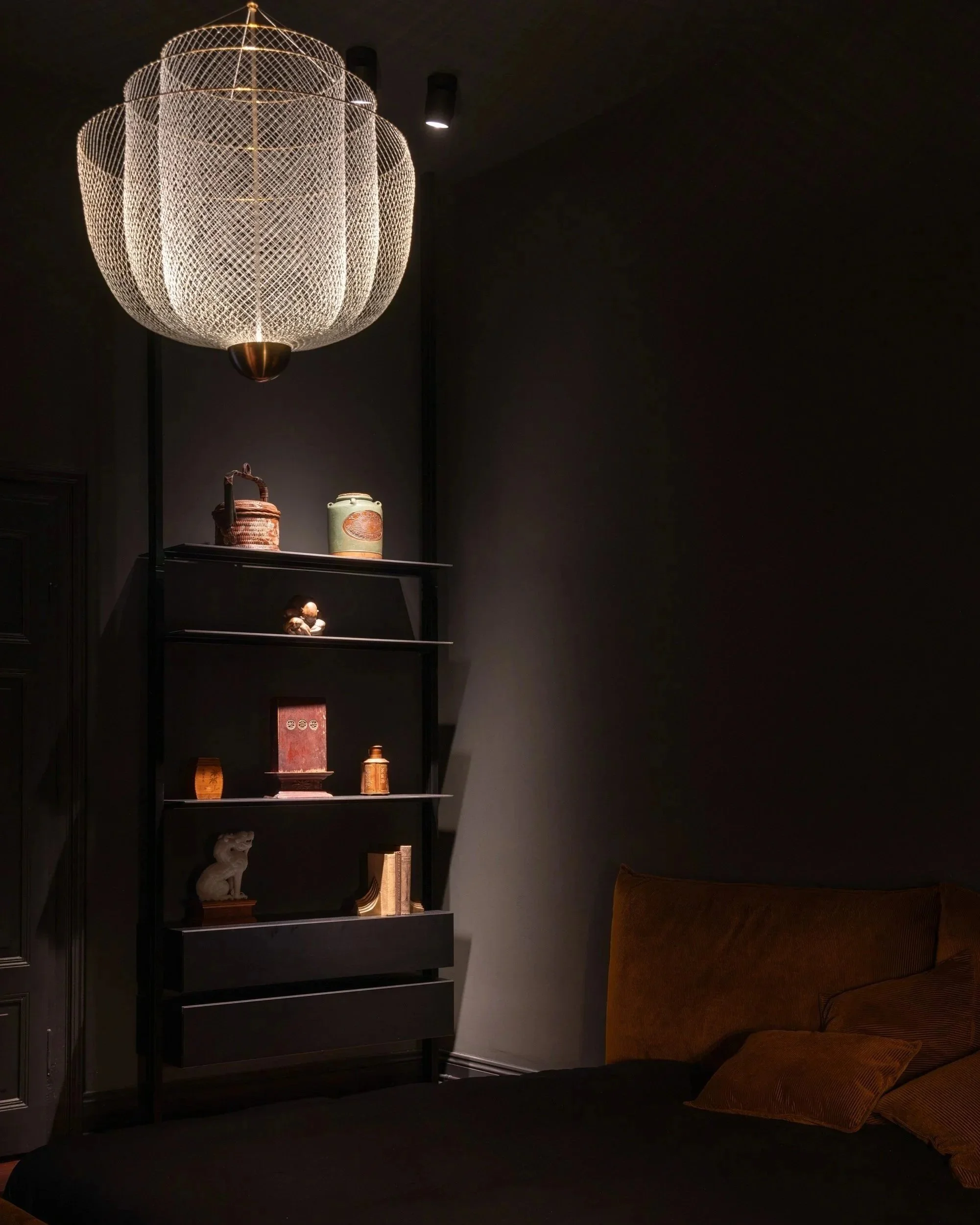 Schwarzer Raum mit braunem Bett, dunkler Wand und einer schwarzen Bücherregal mit dekorativen Gegenständen, Beleuchtung von einer großen, modernen Lampe, gemütliche Atmosphäre.