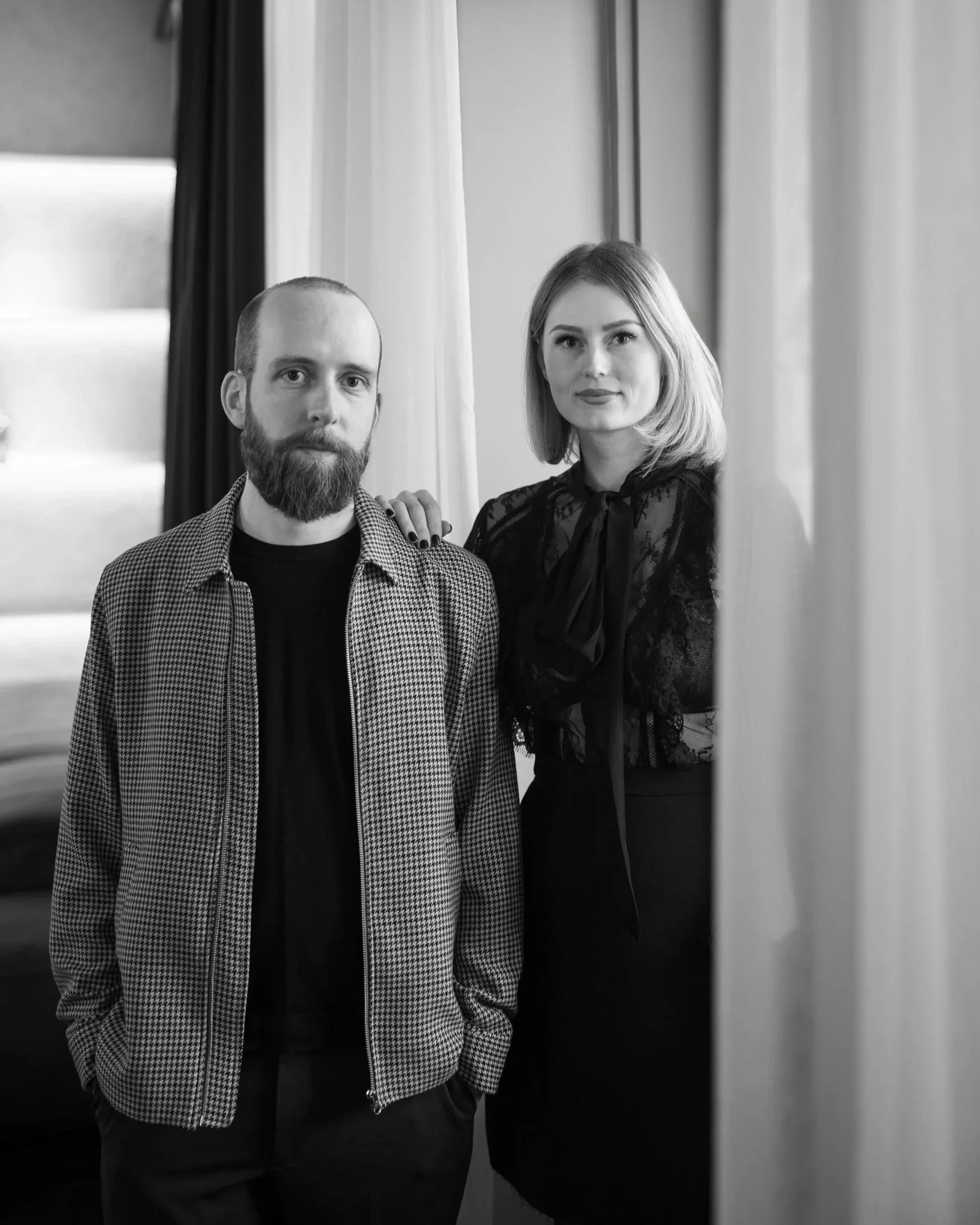 Ein Mann mit Bart und eine Frau mit blonden Haaren posieren im schwarzen und weißen Foto vor Vorhängen.