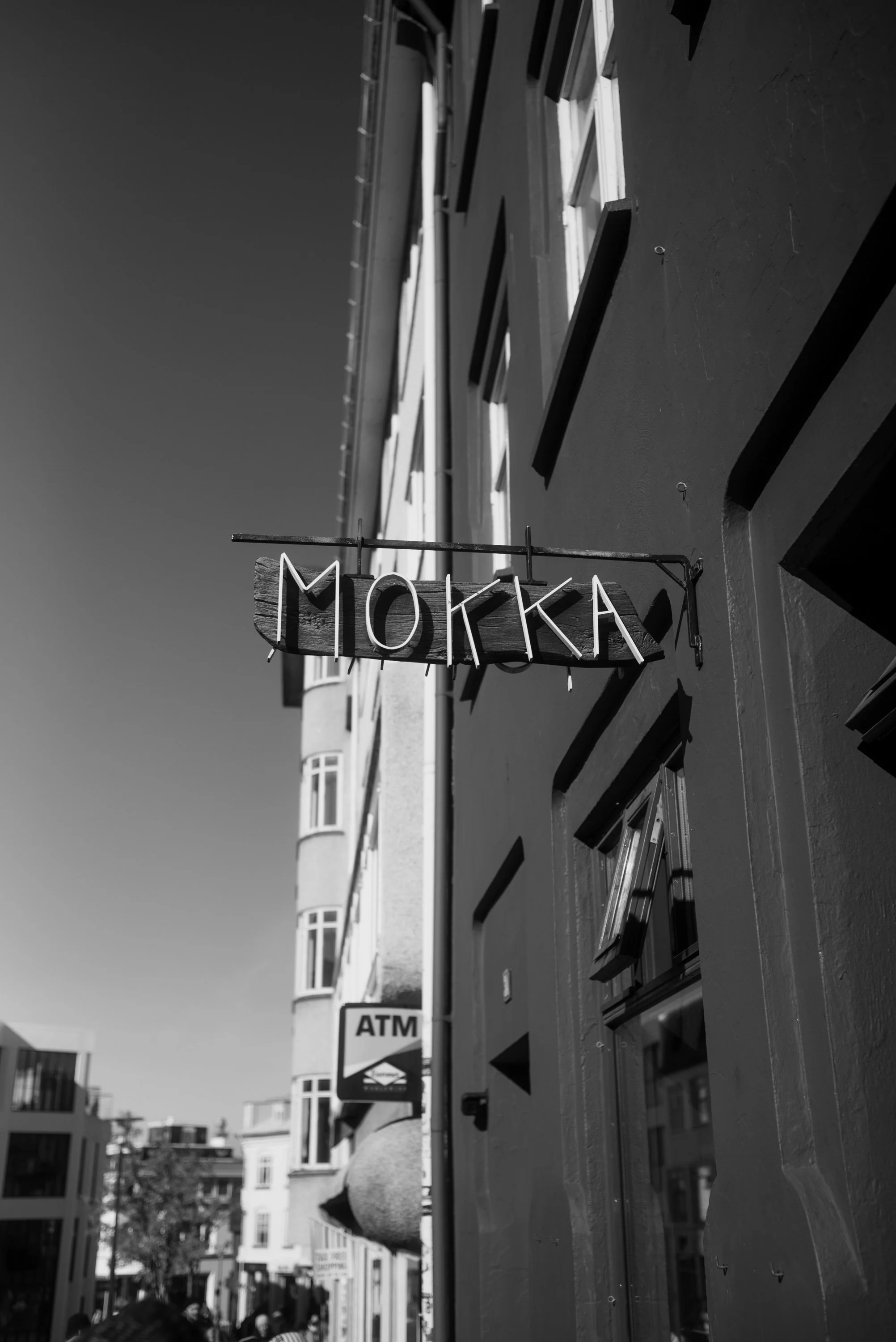 Schwarze und weiße Straßenszene mit Ladenfront, einem leuchtenden Lichtzeichen über der Tür mit dem Wort "MOKKA" und einem ATM-Geldautomaten im Hintergrund.