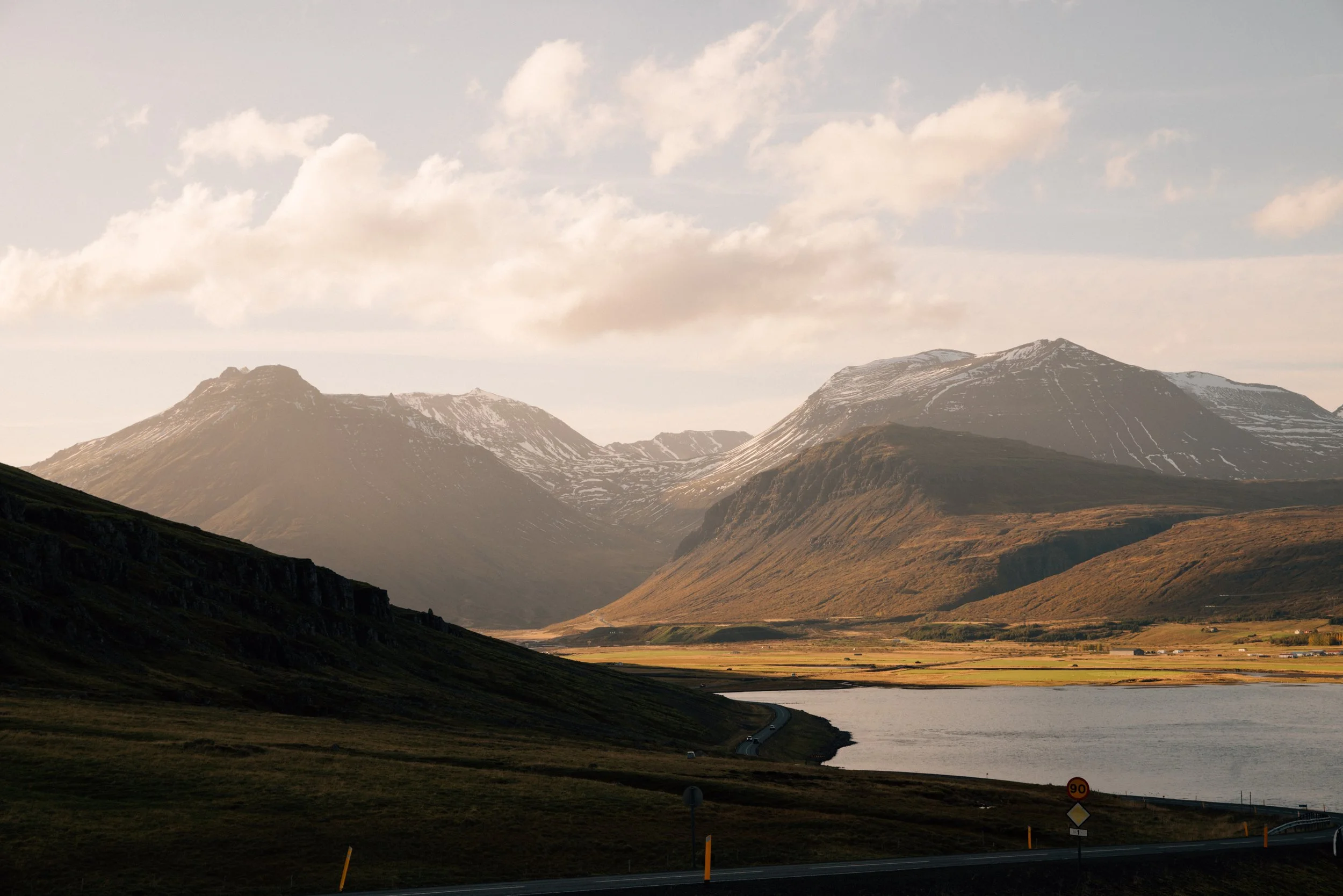 Eine Landschaft mit Bergketten, teilweise schneebedeckten Gipfeln, einem Fluss und einer Straße im Vordergrund, bei Sonnenuntergang oder -aufgang