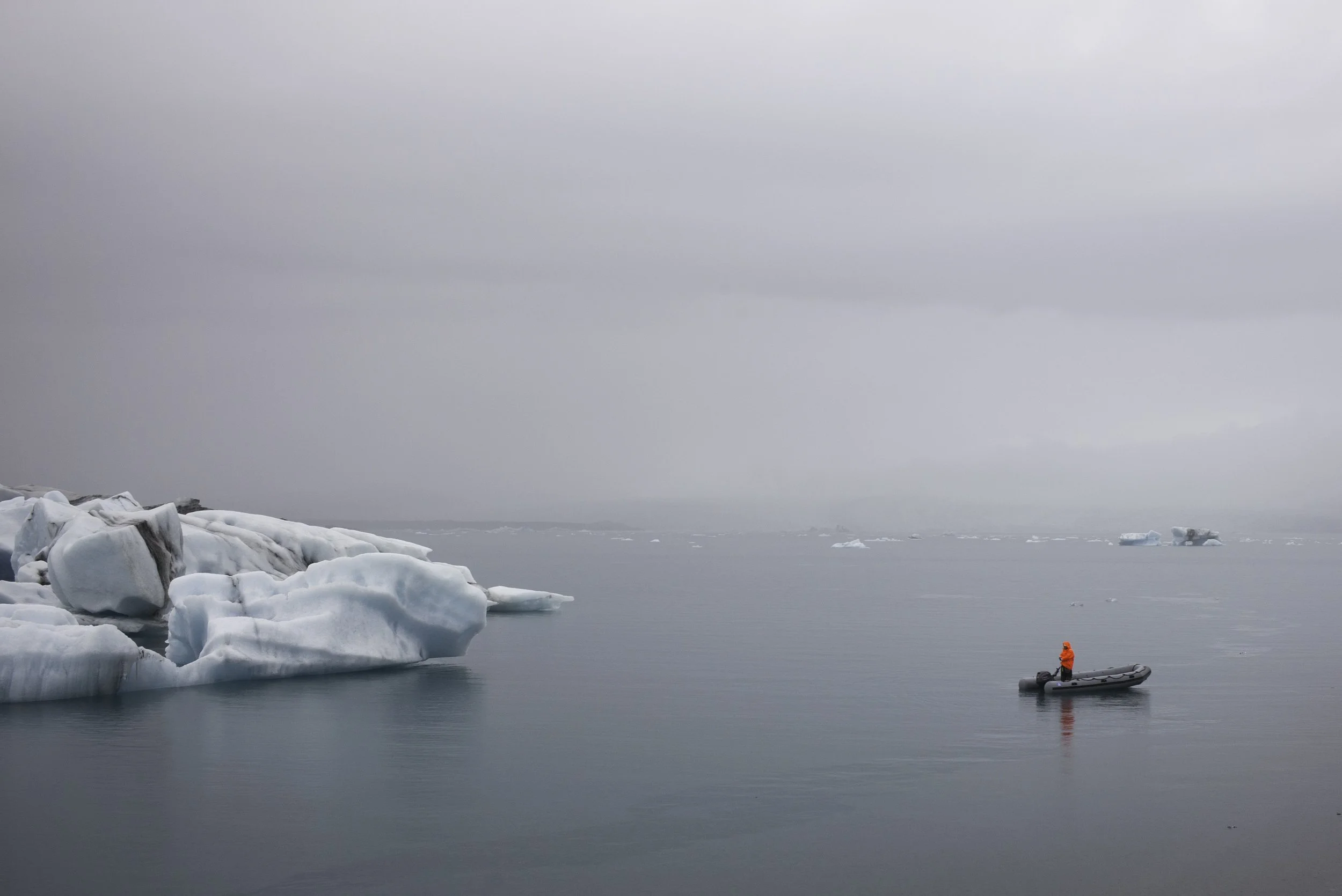 Eine Person in einem kleinen Boot trägt eine orange Jacke in einem polaren Gewässer mit Eisbergen und der Himmel ist bedeckt.