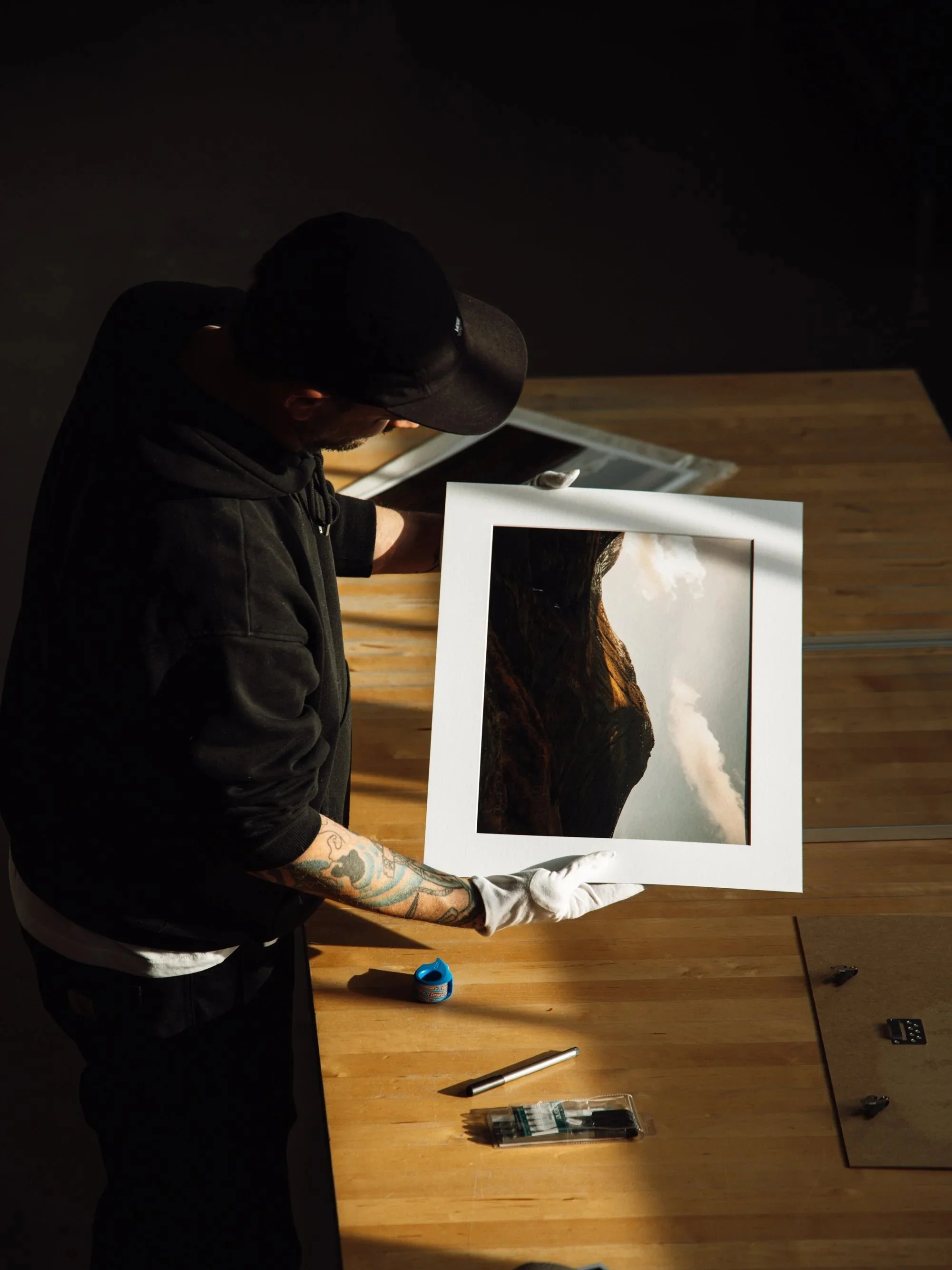Ein Mann in dunkler Kleidung und Baseballkappe hält ein großes fotografiertes Bild eines Bergpanoramas. Er steht an einem Holztisch mit verschiedenen Werkzeugen und leuchtet auf das Bild.