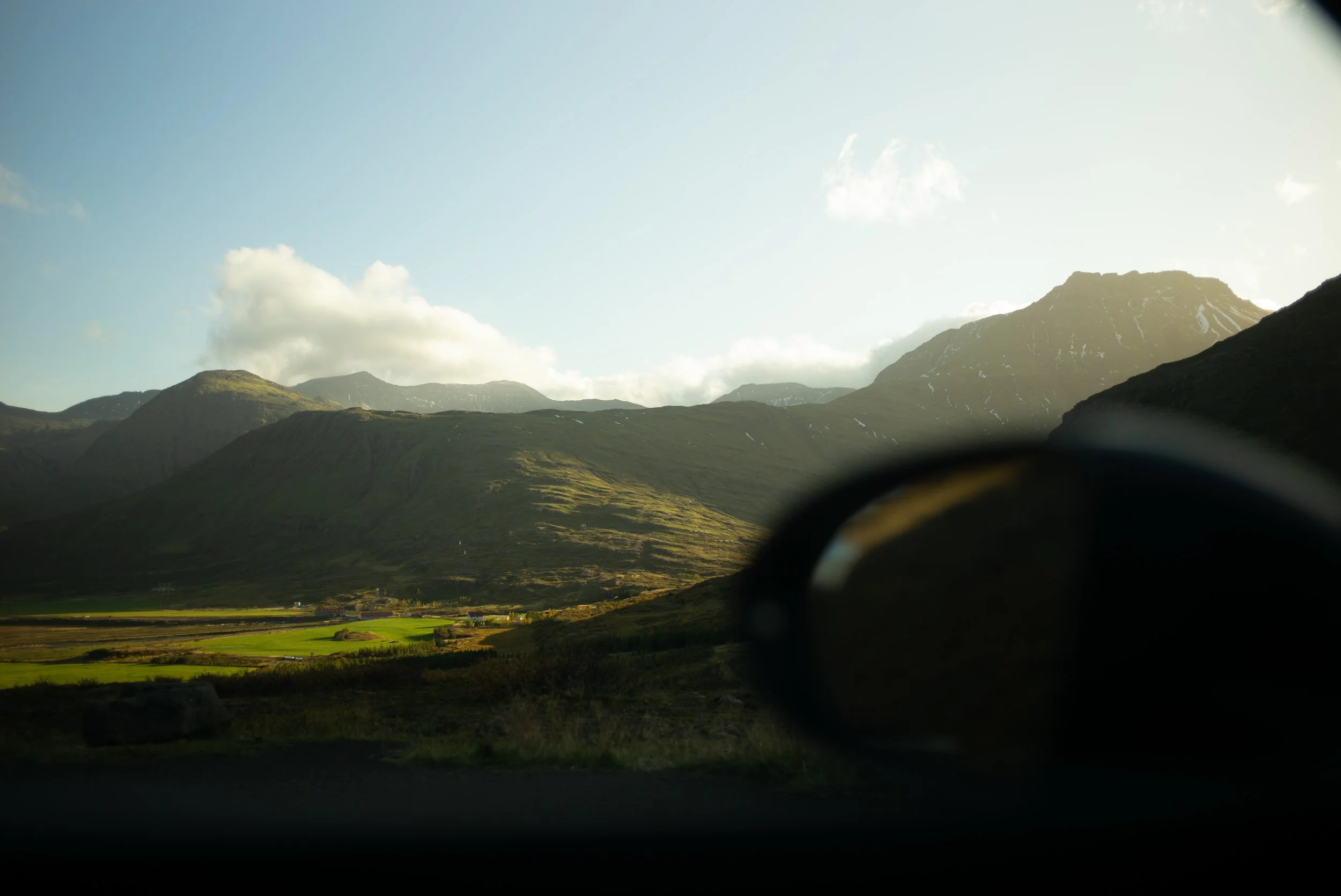 Landschaft mit Bergen, grüne Wiesen und Himmel mit Wolken, Blick durch ein Autospiegel im Vordergrund.