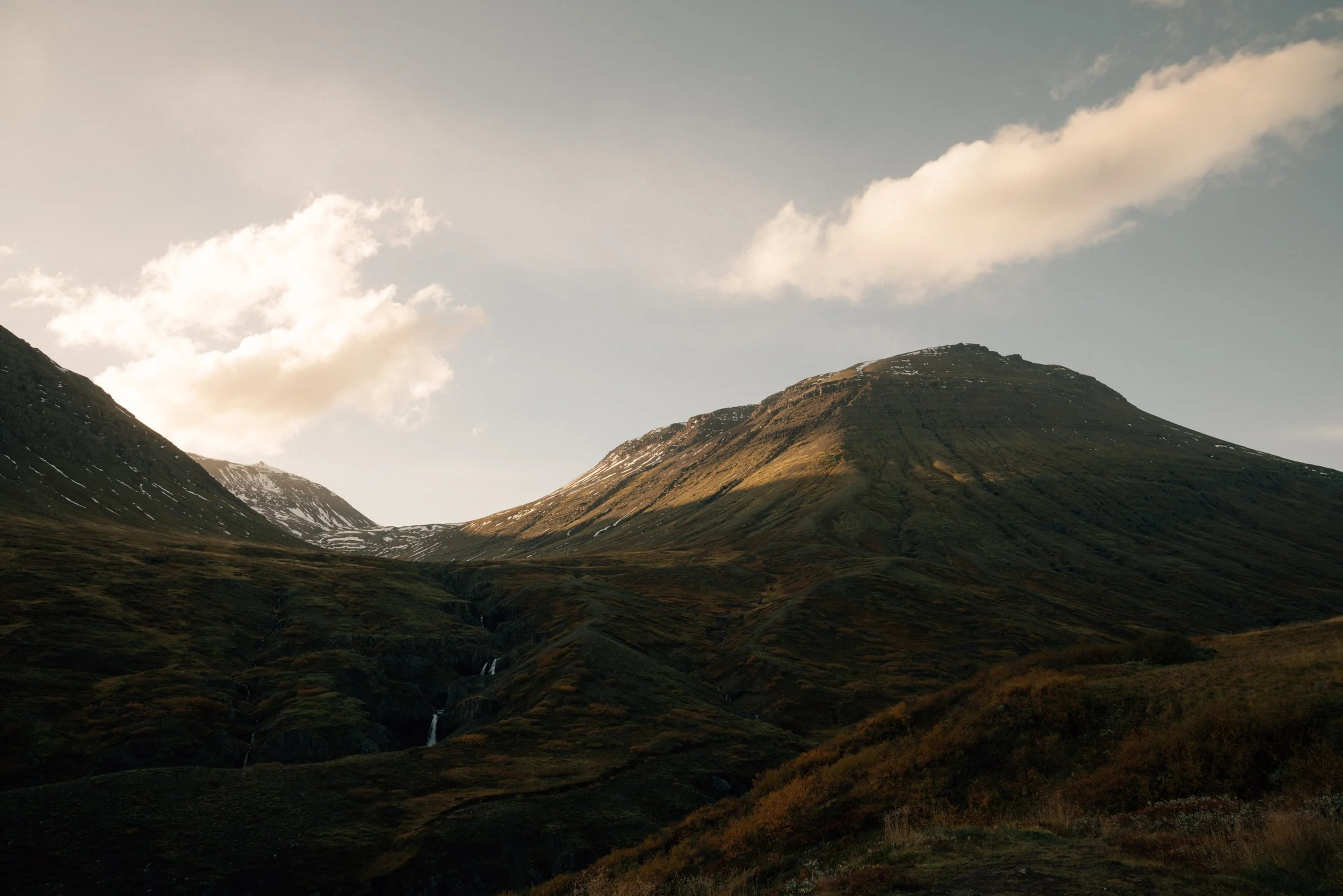 Berglandschaft mit Hügeln und Wasserfällen unter einem bewölkten Himmel.