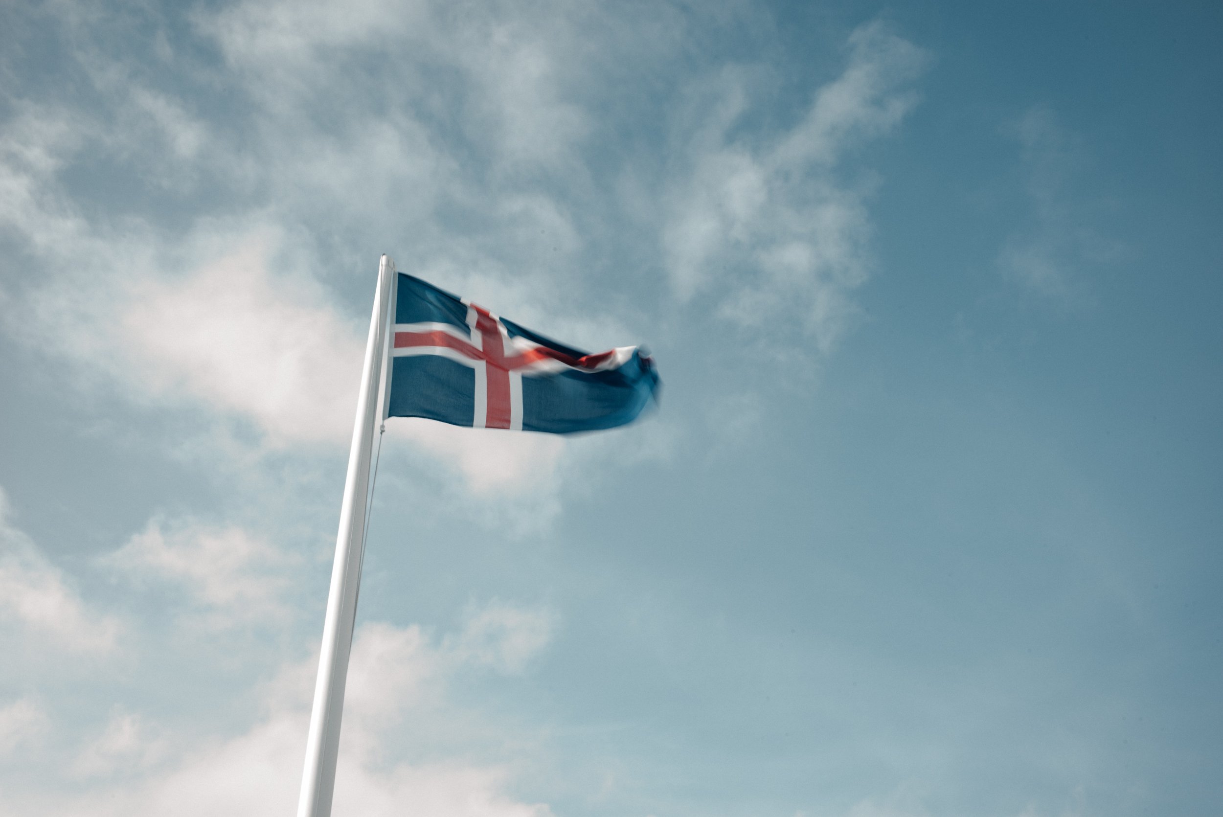 Eine Flagge mit dem design der isländischen Flagge weht im Wind gegen einen blauen Himmel mit wenigen Wolken.