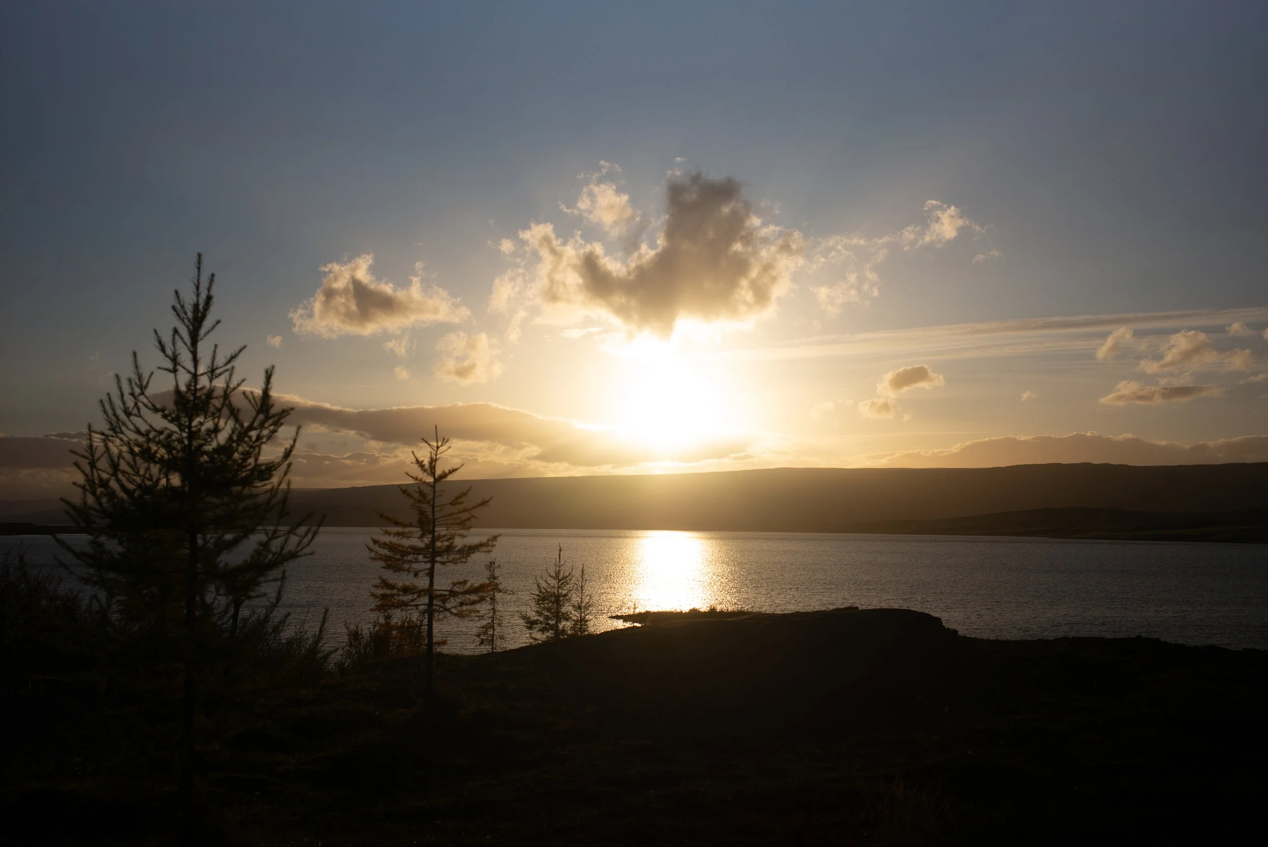 Sonnenuntergang über einem See mit Bäumen im Vordergrund und Hügeln im Hintergrund.