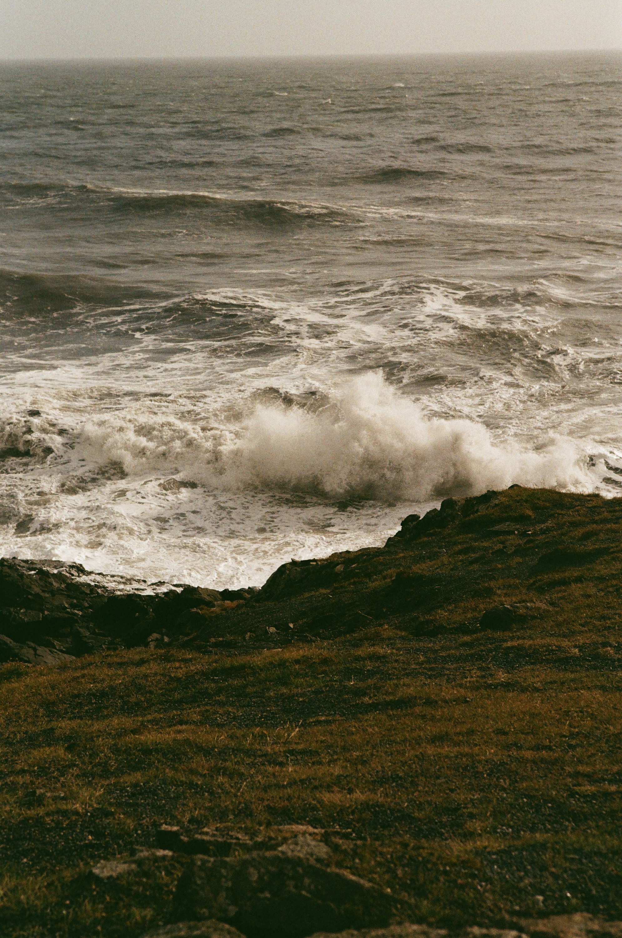 Aufnahme von stürmischer See mit Wellen, die gegen Klippen schlagen, unter bewölktem Himmel.