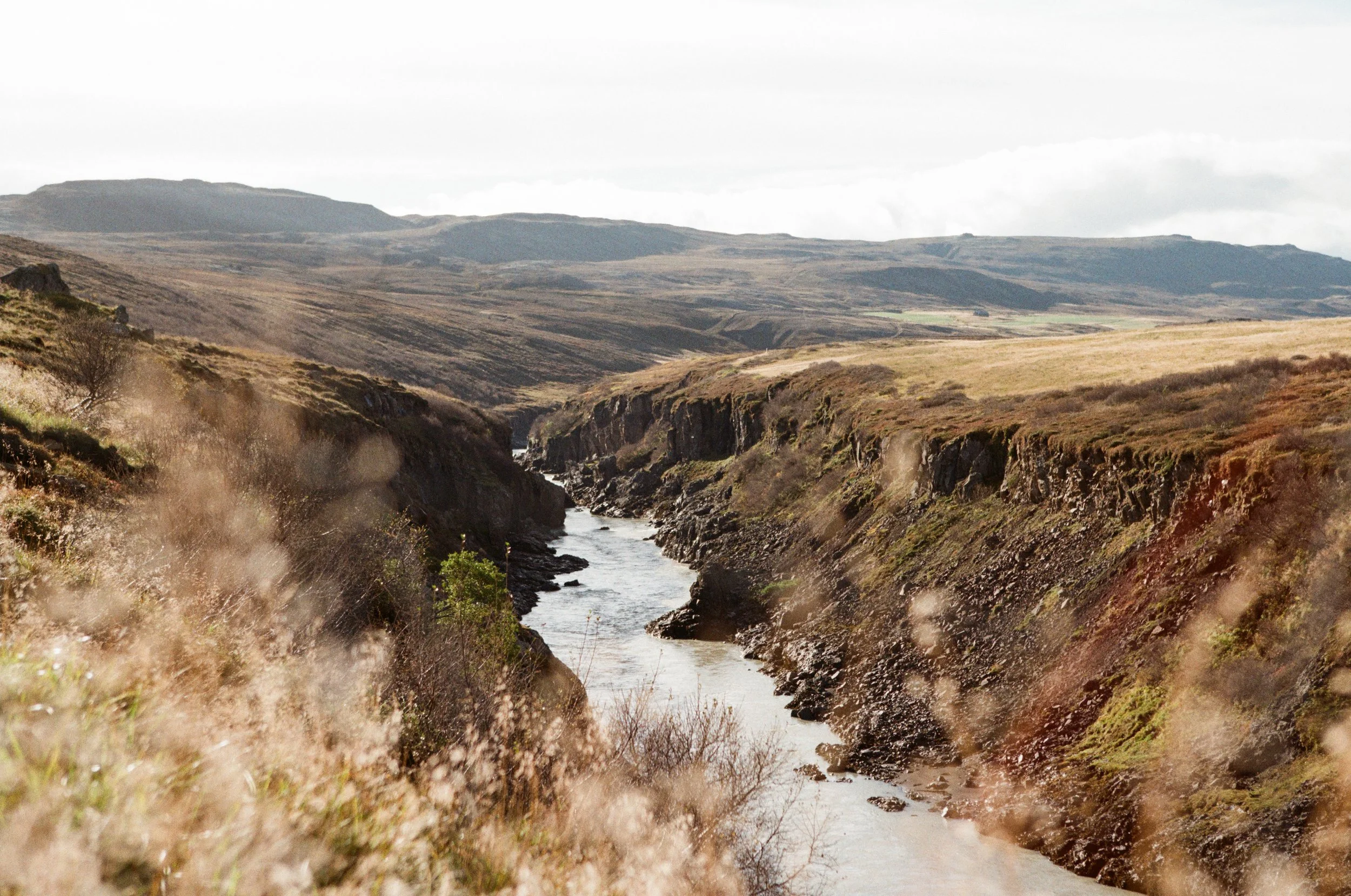 Eine Fluss gesäumt von Steilklippen in einer offenen Landschaft mit karger Vegetation, Berge im Hintergrund, leicht bewölkter Himmel.
