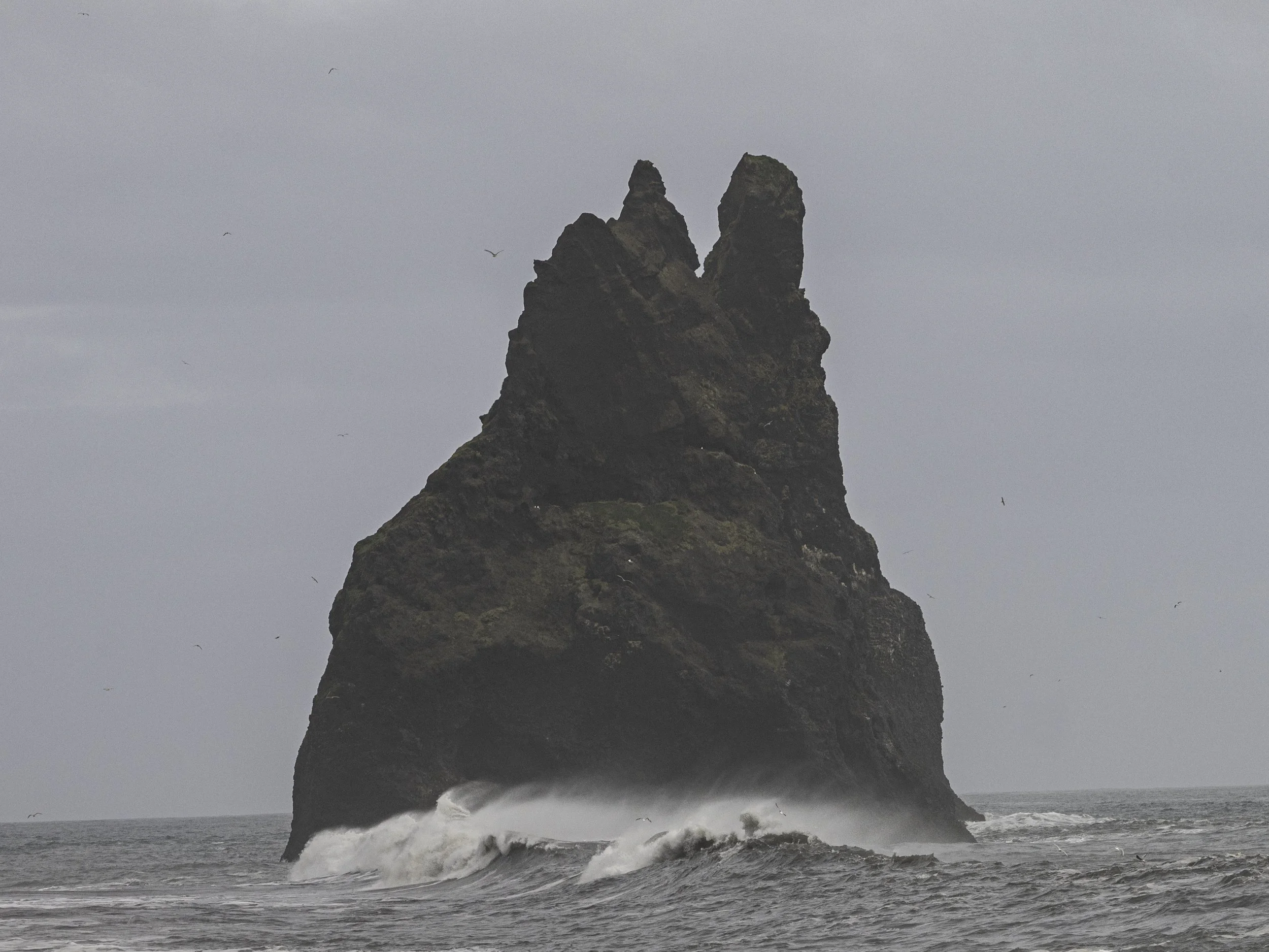 Ein großer, dunkler Felsen im Meer, umgeben von Wellen, bei bewölktem Himmel.