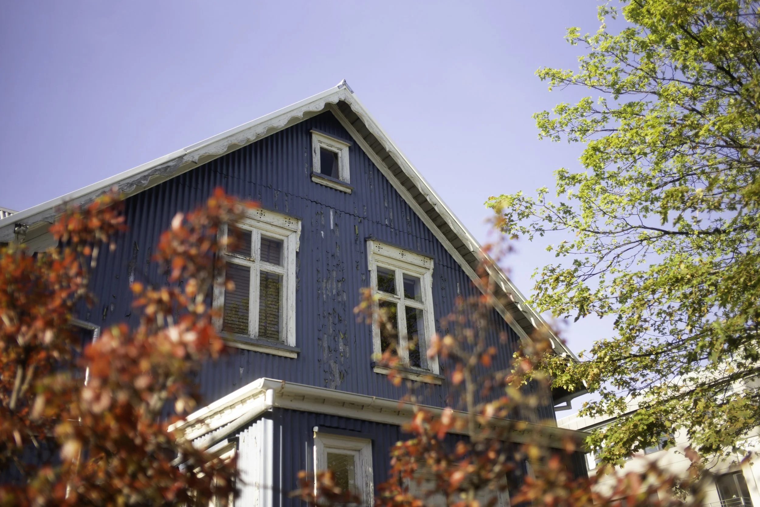 Oberer Teil eines blauen, älteren Hauses mit weißen Fensterrahmen, umgeben von Bäumen mit roten und grünen Blättern, bei sonnigem Himmel.