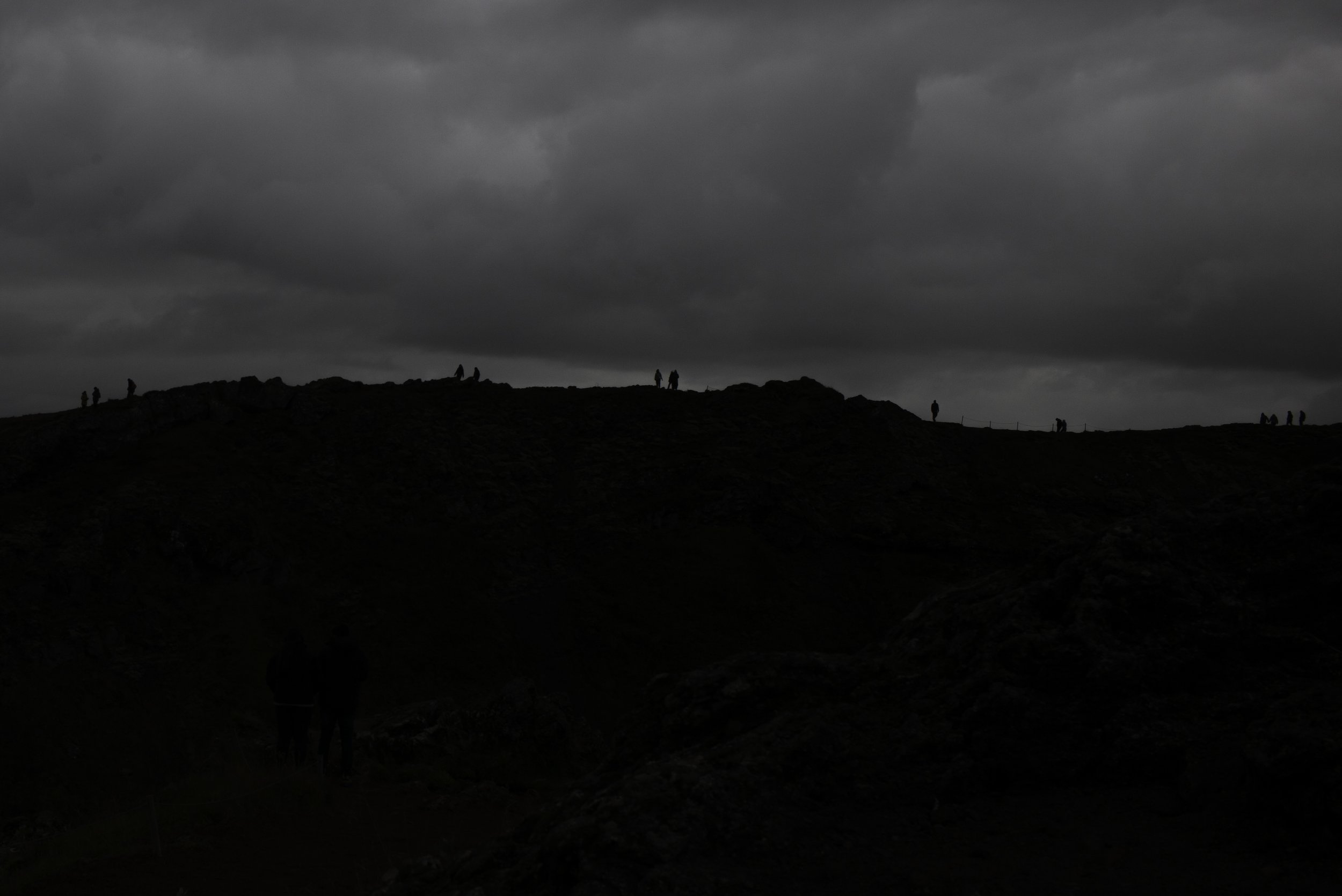 Siluetten von Menschen auf einem Hügel bei dunklem Himmel mit Wolken, im Vordergrund ist die Dunkelheit, im Hintergrund der Himmel grau und bewölkt.