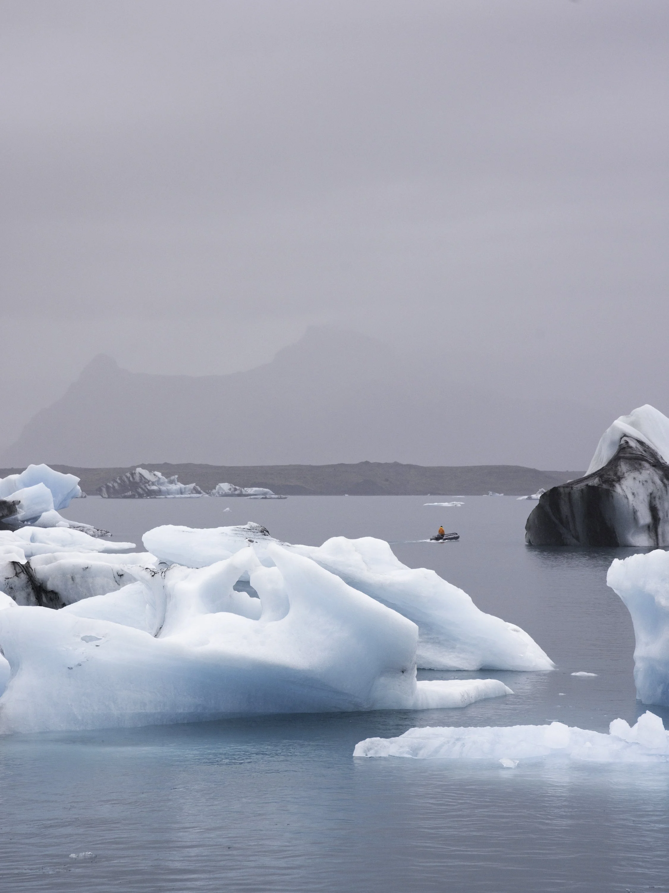 Eisberge im Wasser mit einem kleinen Boot und einem Menschen in oranger Kleidung, im Hintergrund Berge und eine bewölkte Himmel