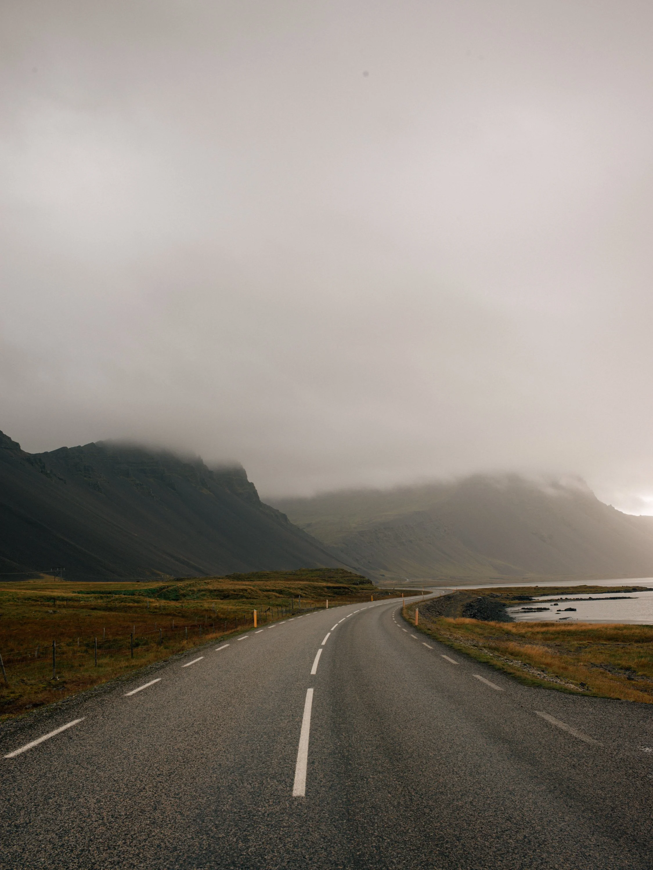 Eine kurvige Straße führt durch neblige Berge und an einem See entlang, mit grauem Himmel.
