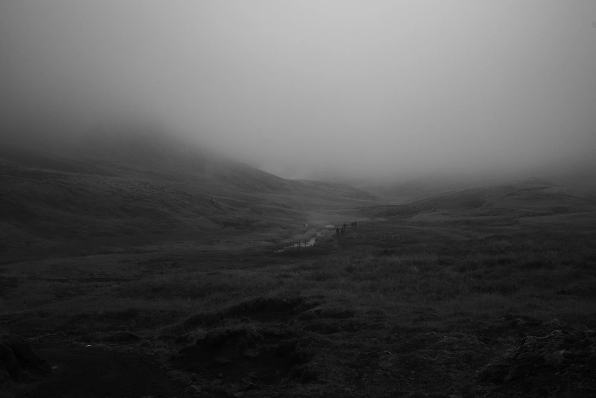 Neblige Hügellandschaft mit einem kleinen Fluss und wenigen Wanderern in der Ferne, schwarz-weiß.