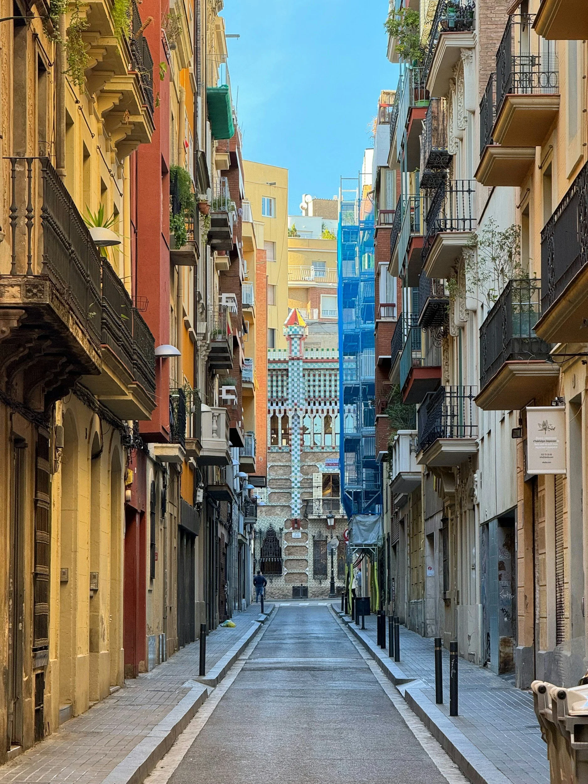 A narrow city street lined with tall buildings on both sides, with balconies and plants. In the distance, there is a colorful, castle-like structure visible at the end of the street under a clear blue sky.