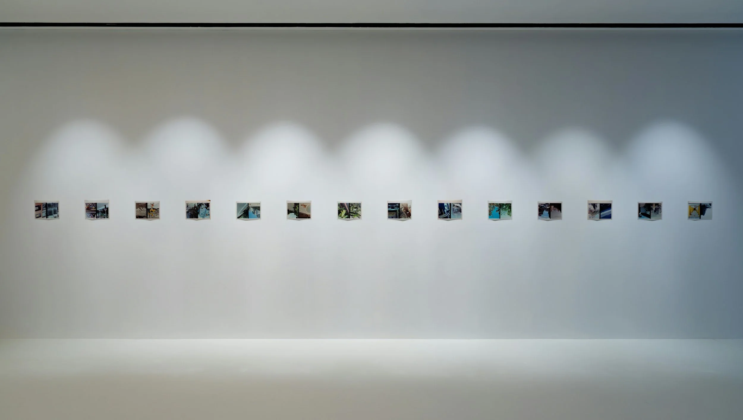 An art gallery wall displaying a horizontal row of small framed photographs arranged evenly. The photographs depict various indoor and outdoor scenes, including trees, buildings, and interiors. The minimalistic white wall and soft lighting highlight the artwork.