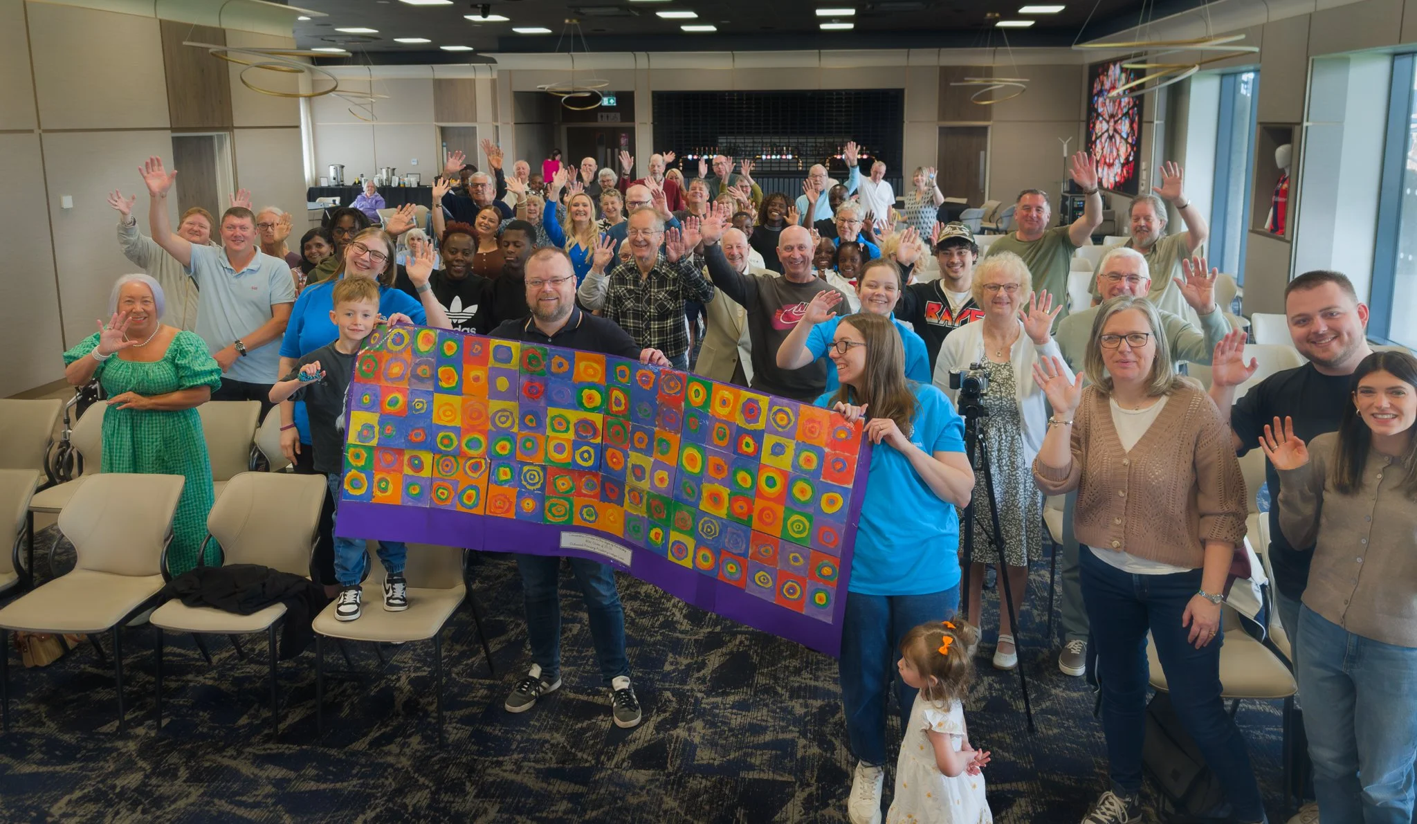 Group of diverse people celebrating at an indoor event, holding a colorful painting of abstract circles and squares.
