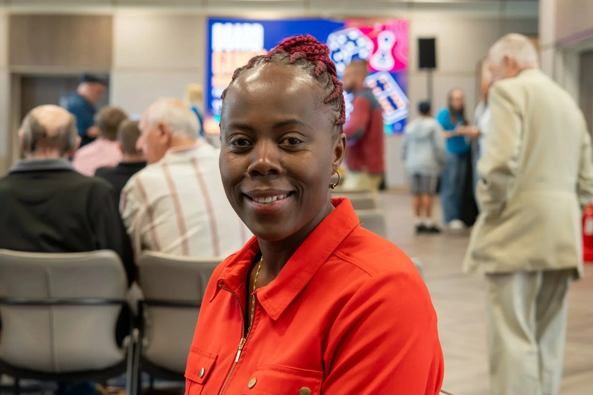 A woman with dark skin and red braided hair, smiling, wearing a red jacket, sitting in a crowded indoor space with chairs, people, and a brightly lit display screen in the background.