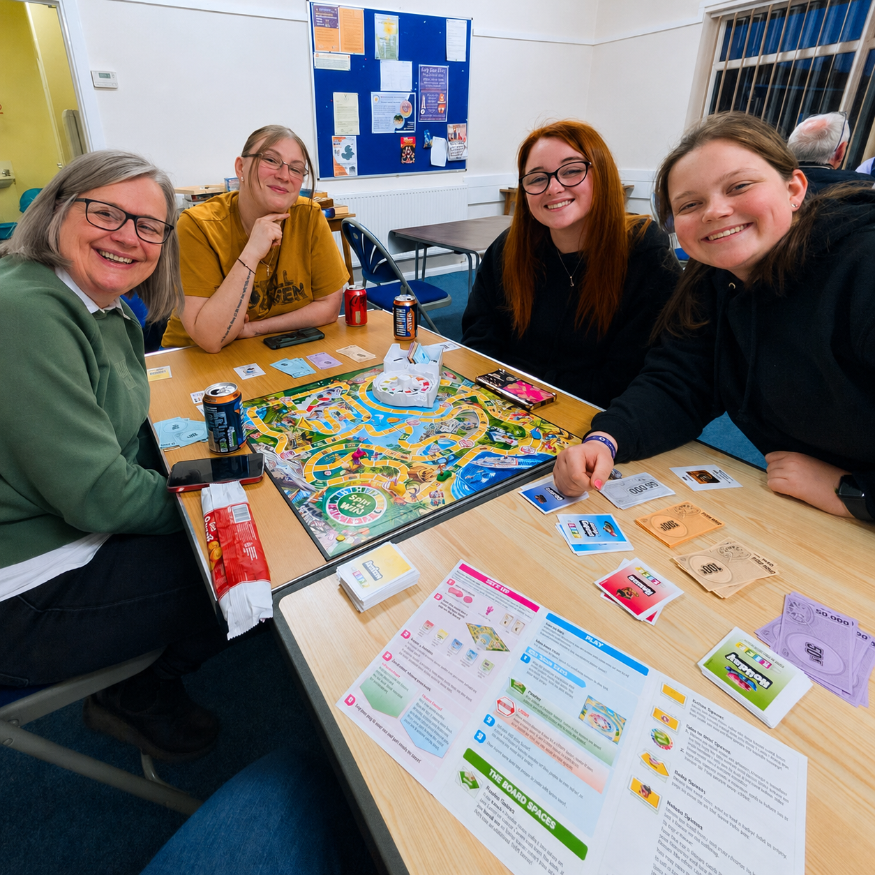 Four women sitting around a table playing Monopoly, smiling at the camera. The table has game pieces, money, and instructional papers. The room has white walls, a bulletin board, and a window with bars.