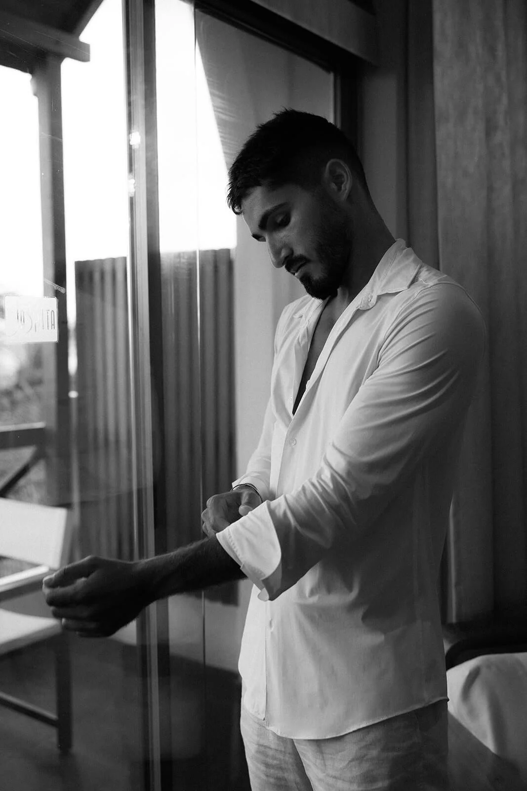 A young man with a beard adjusting his sleeve while looking down, standing near a window with natural light in a room with wooden walls, black and white photograph.