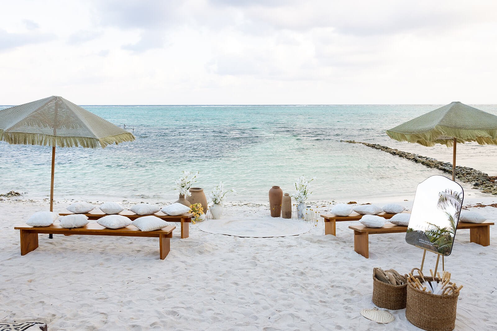 Beach setup with two wooden benches with white cushions and umbrellas, decorative vases and white flowers, mirror reflecting palm trees, and baskets on the sand, overlooking the ocean under cloudy sky.