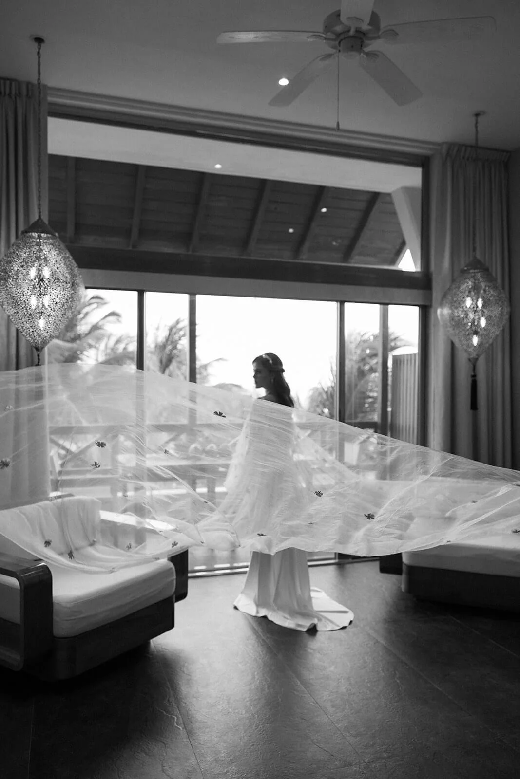 A woman in a wedding dress stands behind a sheer, decorated veil inside a room with large windows and tropical trees visible outside. The room has elegant hanging pendant lights and curtains, with a ceiling fan on the ceiling.