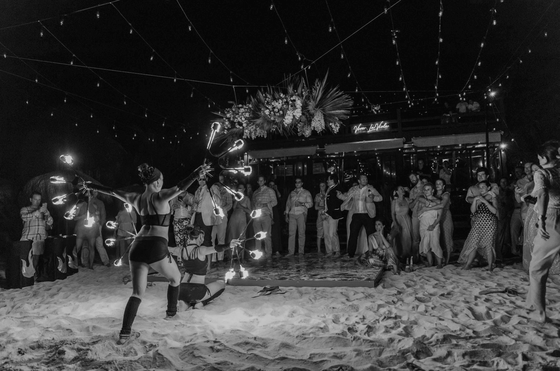 Nighttime beach scene with fire performers and a crowd watching, decorated with string lights and tropical flowers overhead.