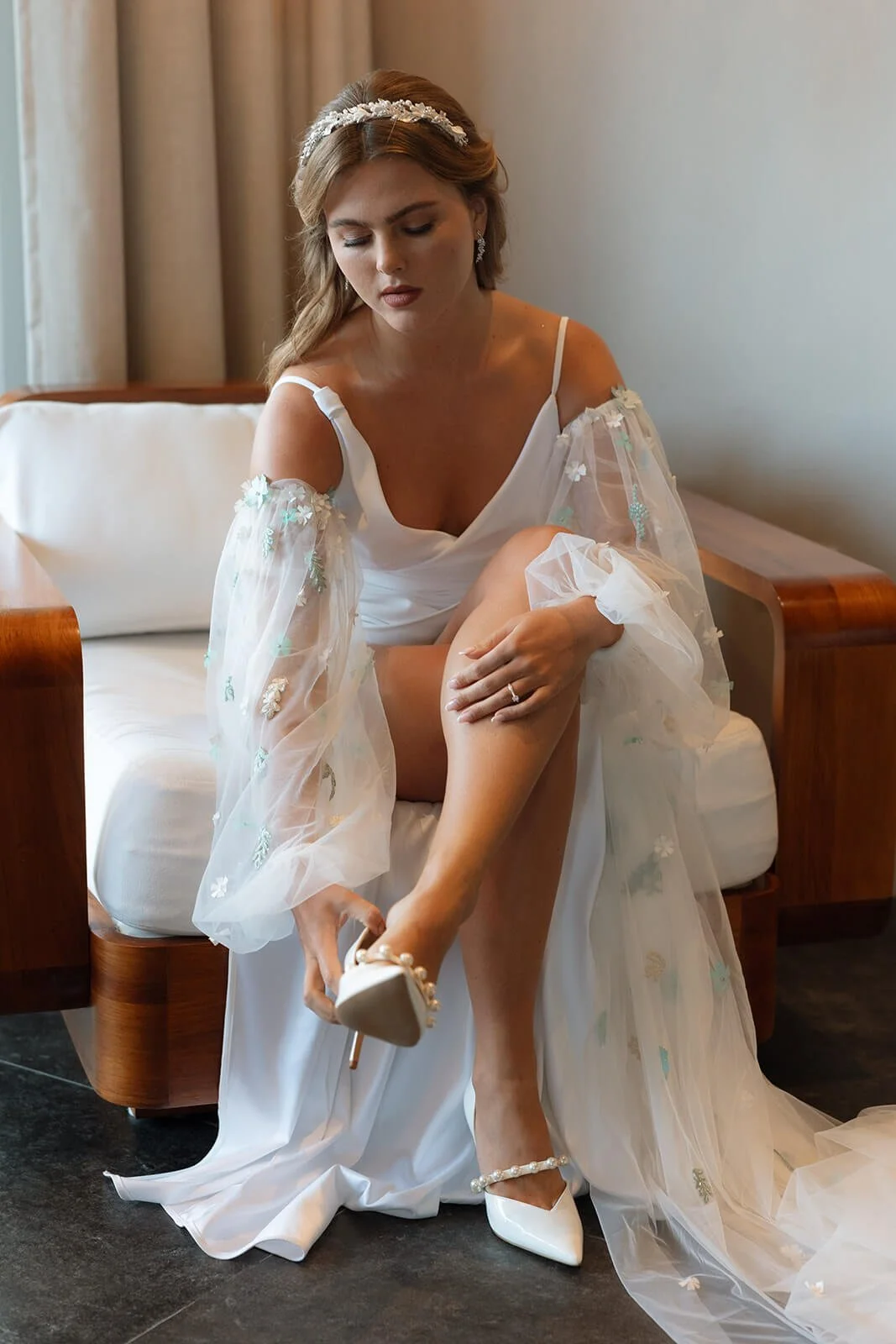 Bride in a white wedding dress sitting on a sofa, adjusting her pearl-embellished white high heel shoe.