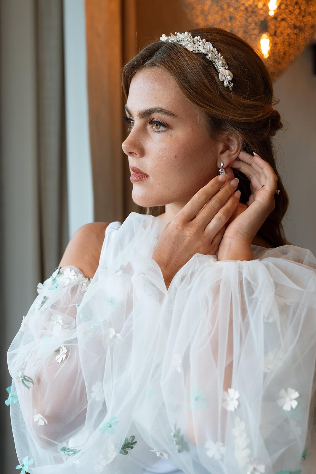 A bride with light brown hair and a floral headband adjusts her earrings, wearing a white dress with off-shoulder floral embroidery, standing near a window with warm lighting.