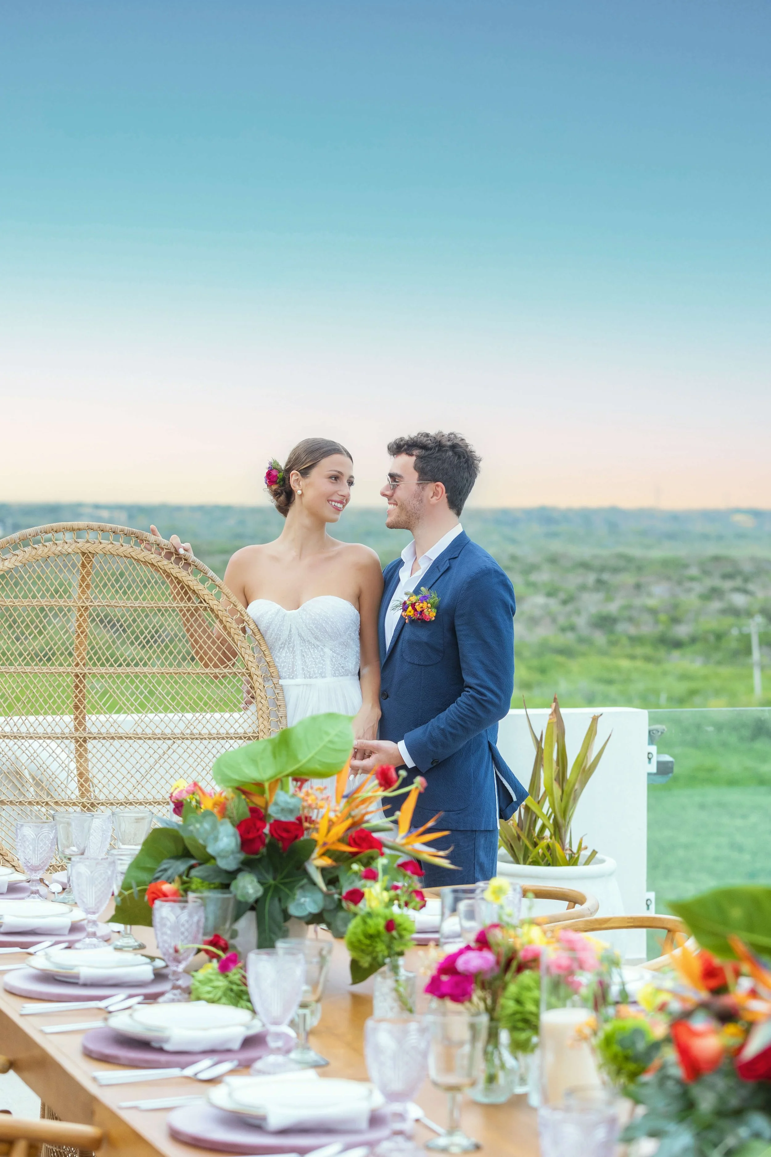 A bride and groom hold hands and smile at each other during a wedding celebration outdoors with a decorated reception table in the foreground and a scenic green landscape in the background.