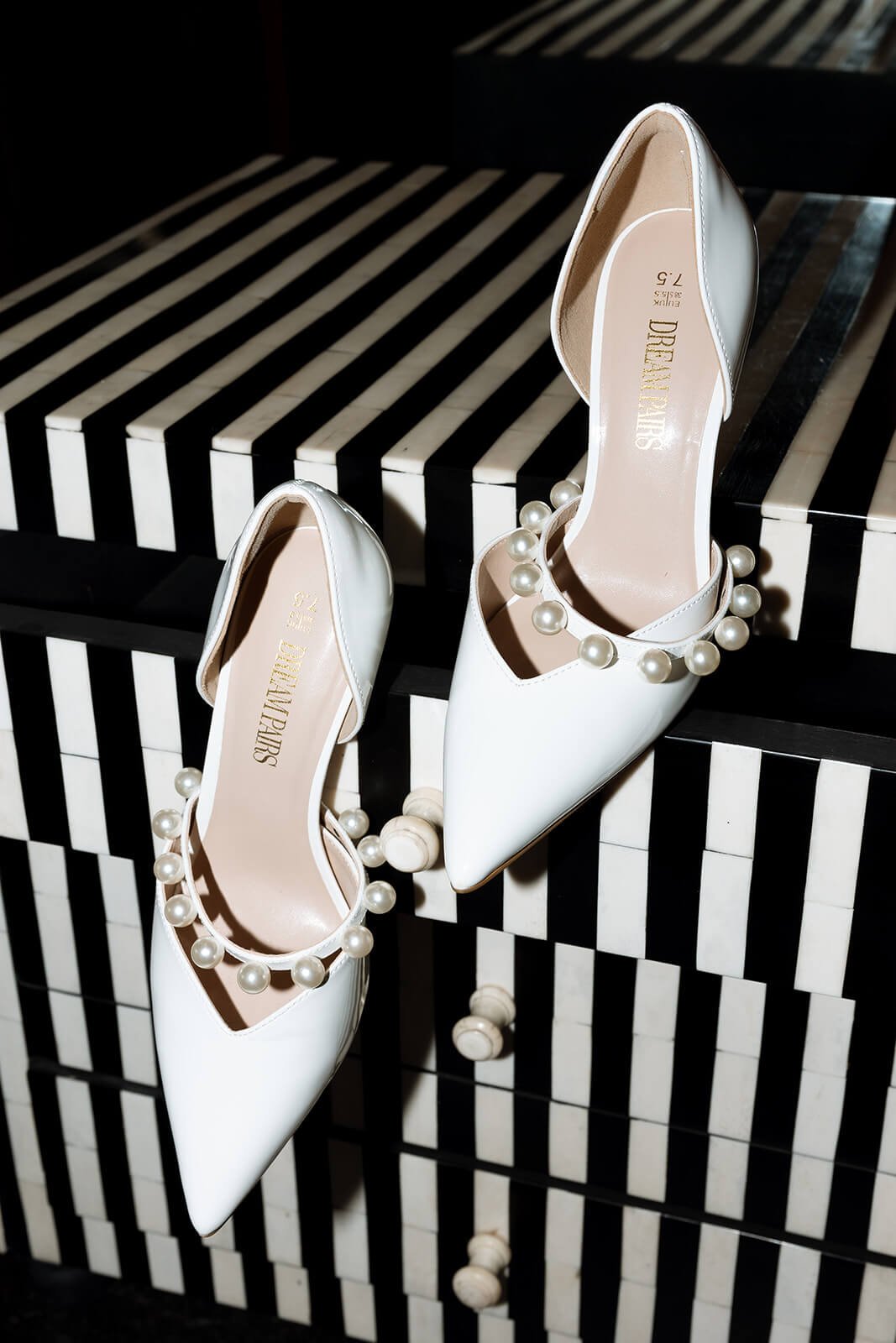 A pair of white high heel shoes with pointed toes and pearl ankle straps, resting on a black and white striped piano.