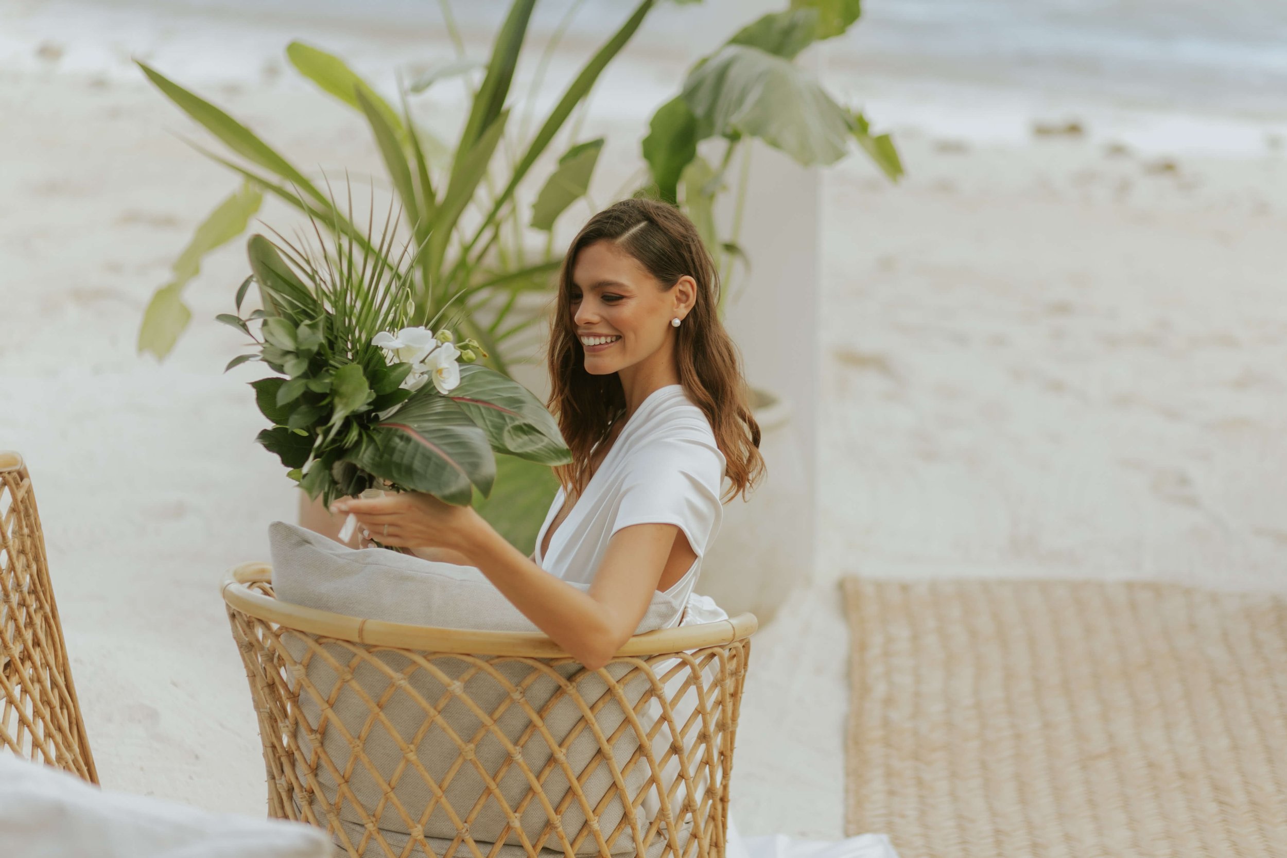 A woman with medium length brown hair smiling while holding a large bouquet of tropical plants with white flowers. She is sitting in a wicker chair with a cushion, in a bright space with green plants and a white brick wall in the background.