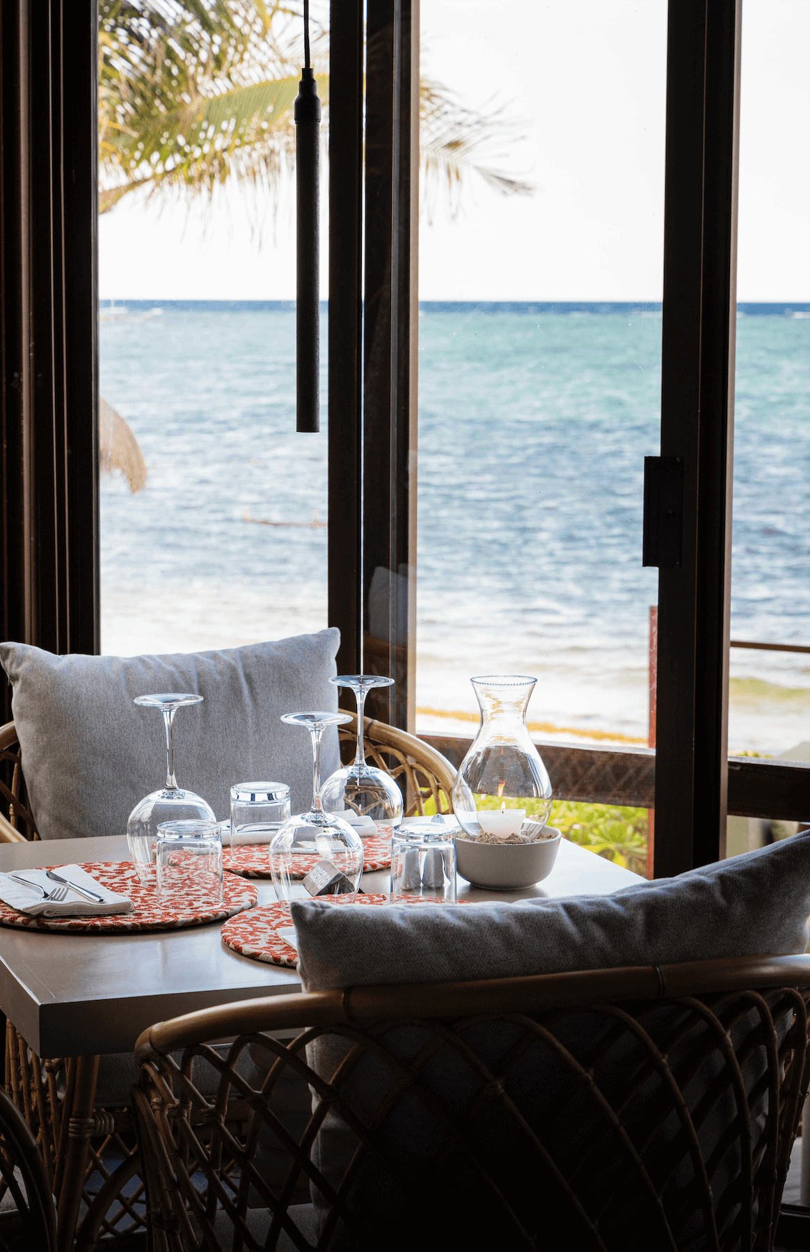 Table set with wine glasses, a water pitcher with a candle inside, and place settings, overlooking a beach and ocean through glass doors.