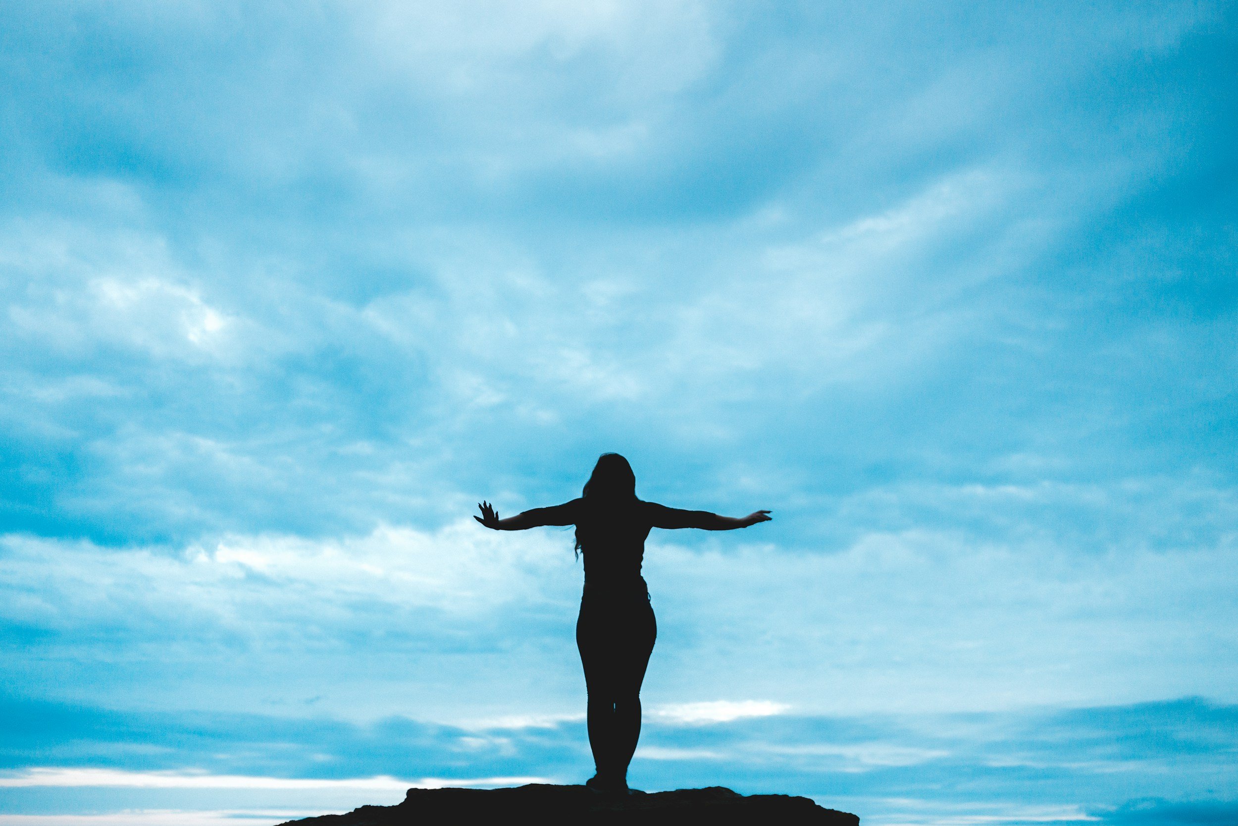 Person standing on a rock with arms outstretched under a cloudy blue sky.