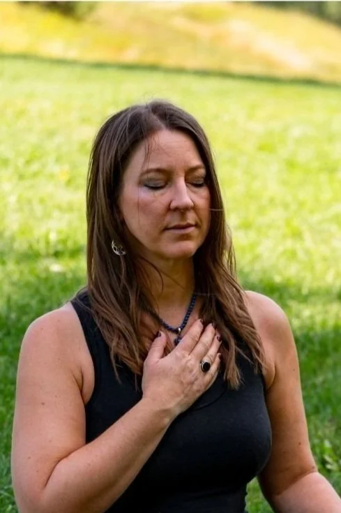 A woman in a black tank top with brown hair and a necklace practicing mindfulness outdoors, with her eyes closed and one hand over her heart.