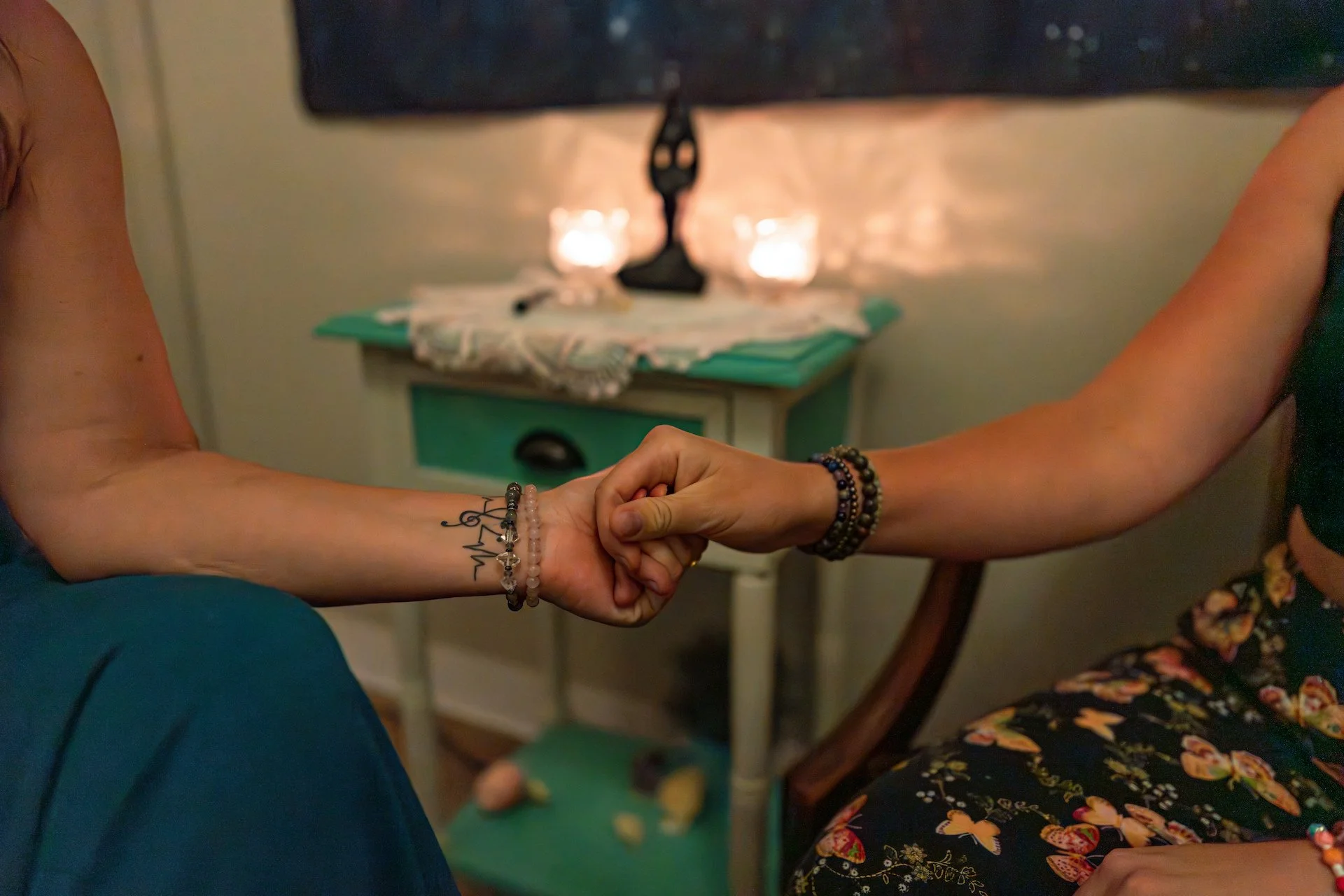 Two people holding hands, with one person showing a tattoo on their wrist, in a cozy room with a small nightstand and lit candles in the background.