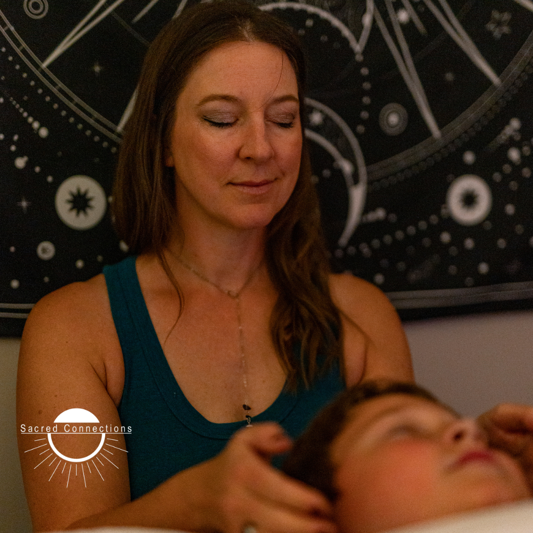 A woman with shoulder-length brown hair, wearing a teal sleeveless top and a necklace, receiving a massage or healing treatment on her head from another person. There is a black cosmic-themed tapestry in the background.